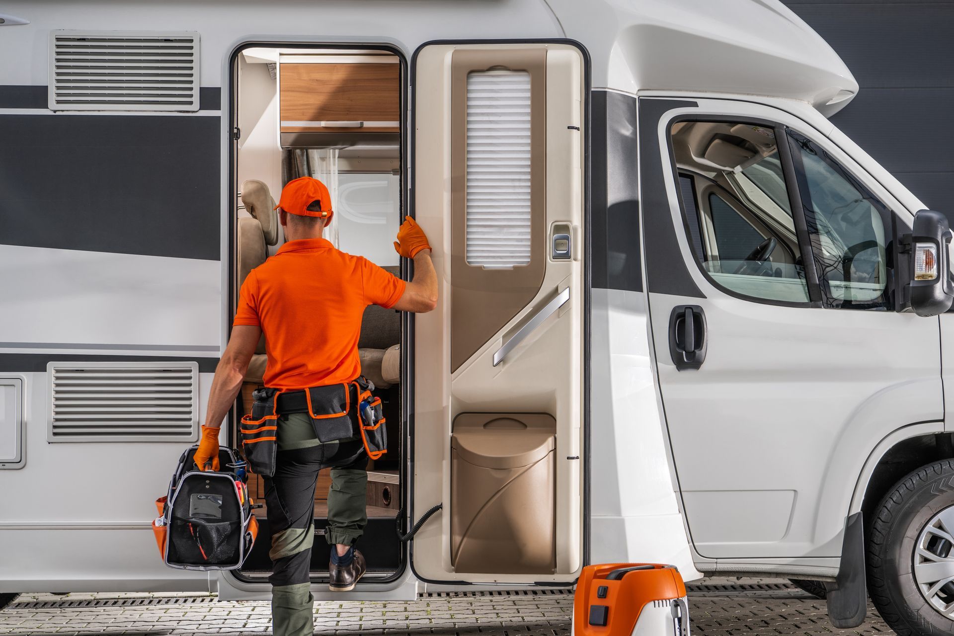 Person in orange shirt and tool belt enters a white RV, holding a toolbox. Person in orange shirt and tool belt enters a white RV, holding a toolbox.