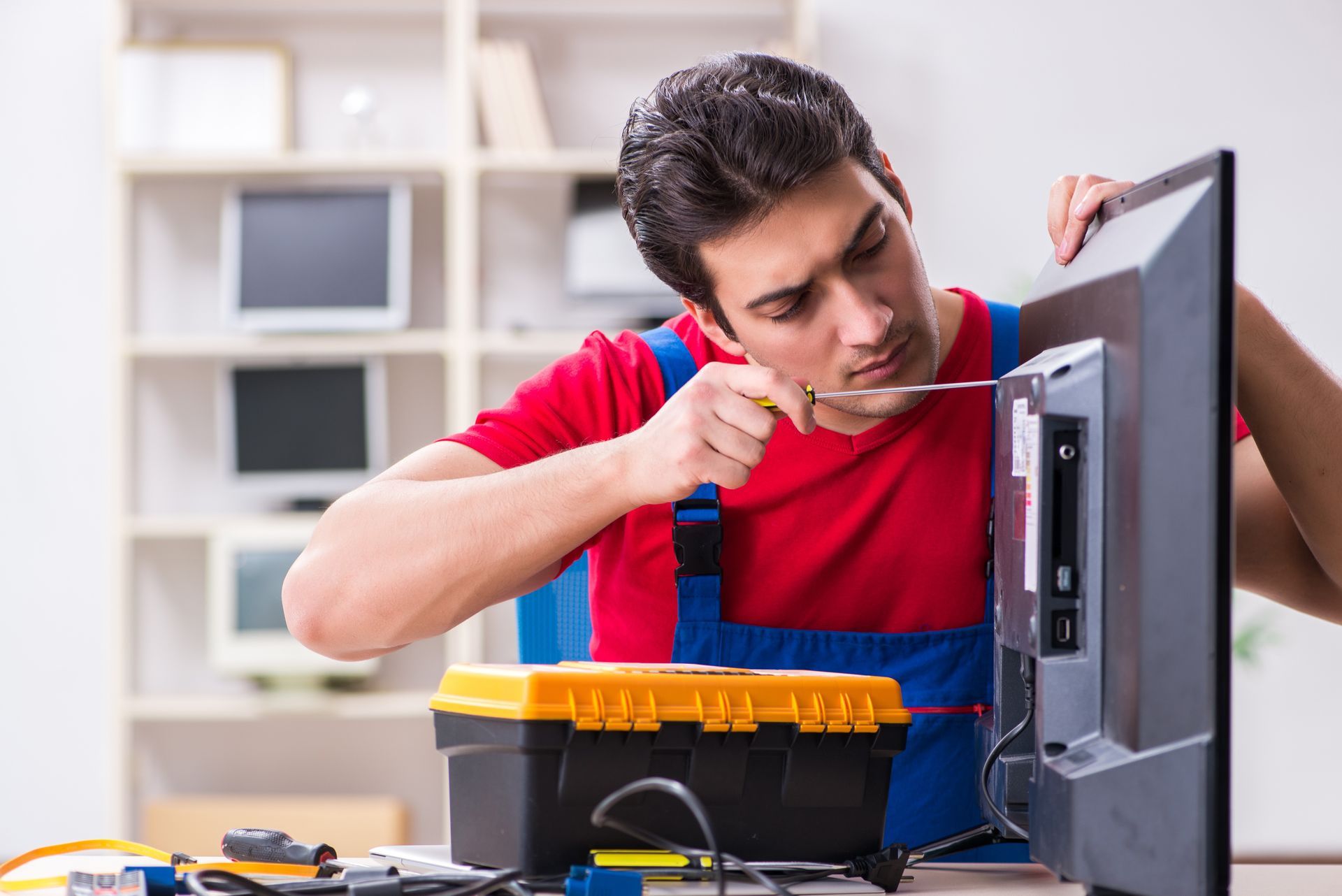 Man in red shirt and blue overalls fixing a TV with a screwdriver in a well-lit room. Man in red shirt and blue overalls fixing a TV with a screwdriver in a well-lit room.