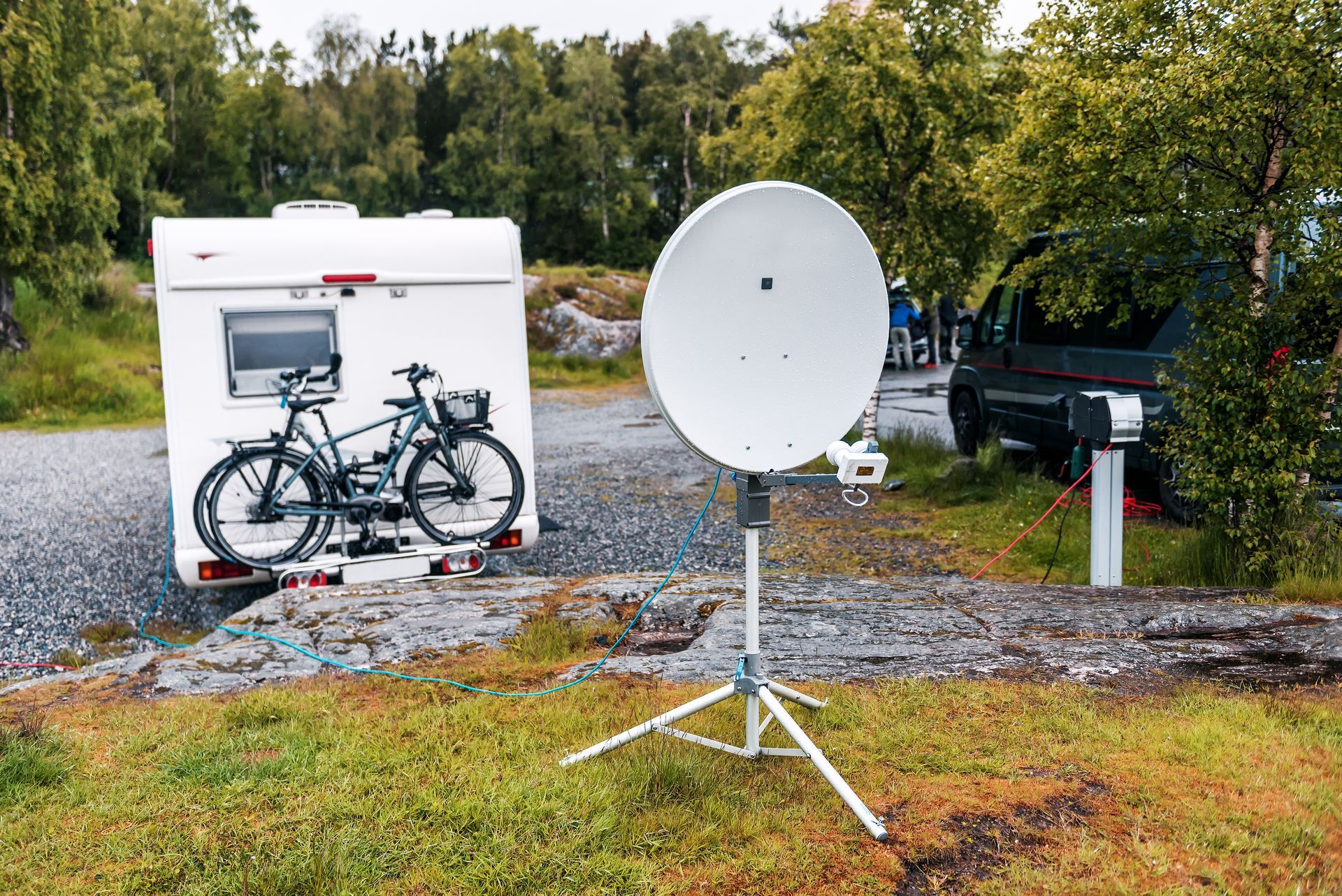 White camper trailer with bikes, satellite dish on tripod, outdoors in wooded area. White camper trailer with bikes, satellite dish on tripod, outdoors in wooded area.