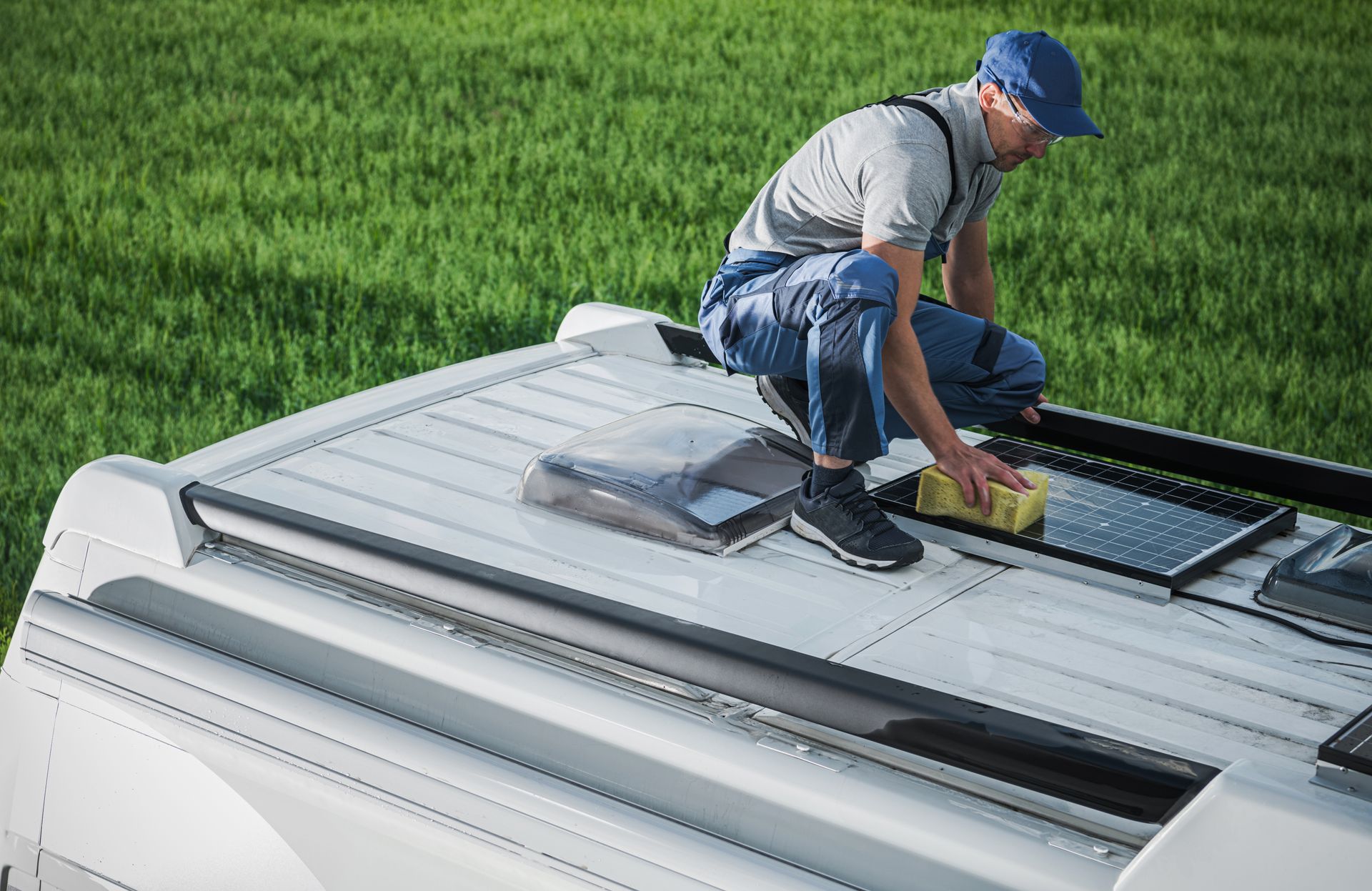 Person cleans a skylight on a white van roof, green field in background.