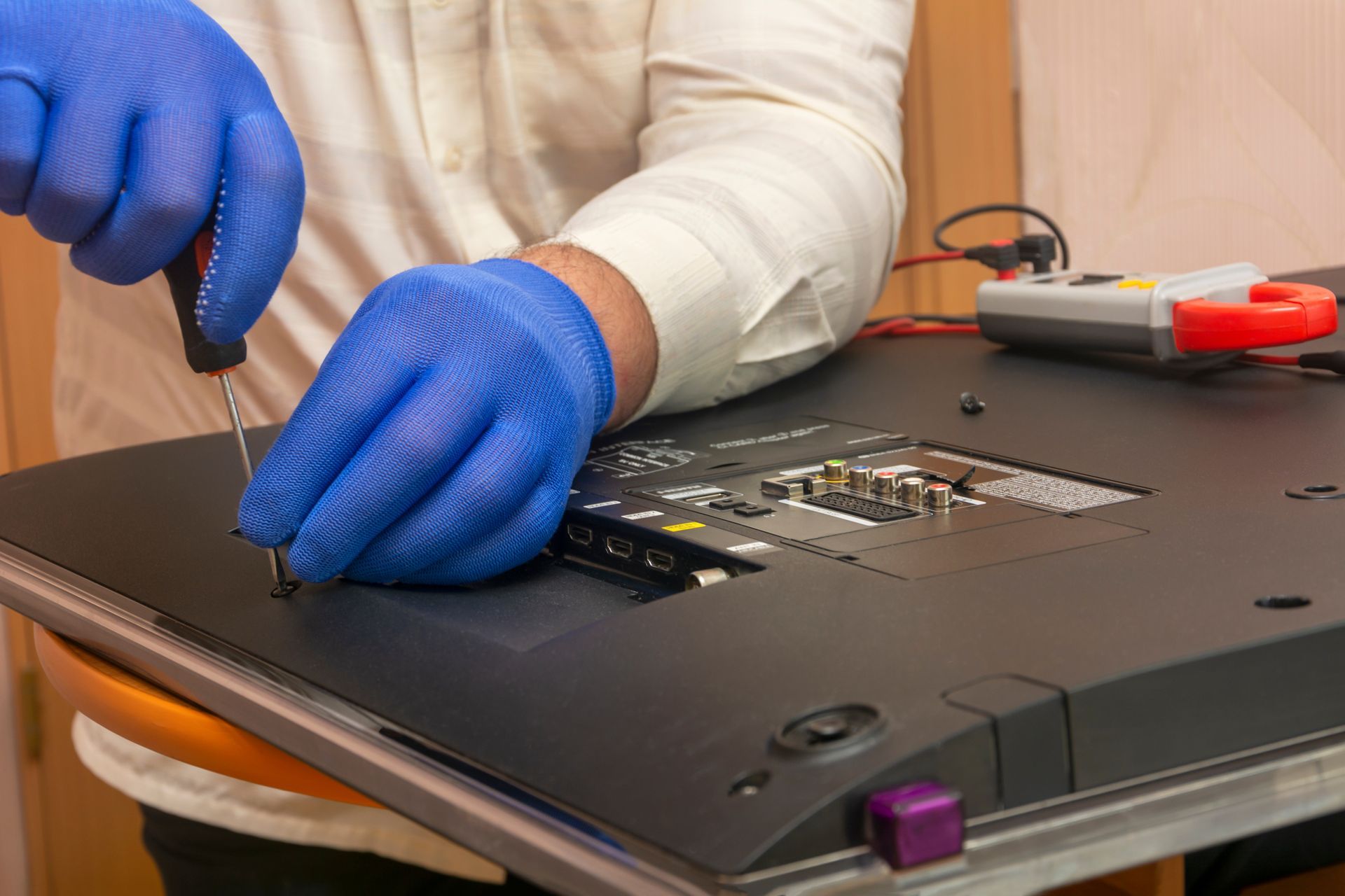 A person wearing blue gloves repairs a TV with a screwdriver, back panel open, in a room. A person wearing blue gloves repairs a TV with a screwdriver, back panel open, in a room.