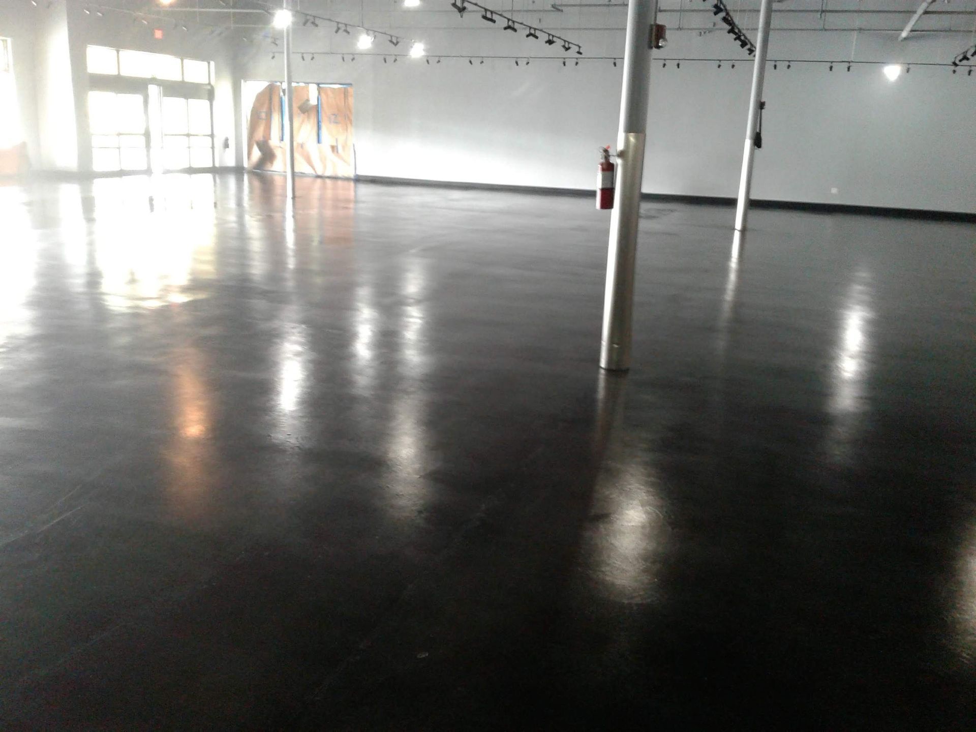 Empty warehouse-like room with glossy black floor, white walls, and bright light from windows.