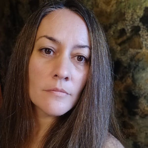 A woman with long hair is standing in front of a stone wall and looking at the camera.