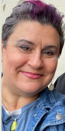 A woman with purple hair and a denim jacket is smiling for the camera.