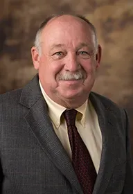 Smiling man in a gray suit and burgundy tie against a blurred brown studio background