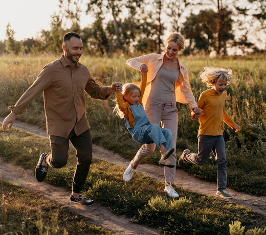 Family running hand-in-hand on a sunlit dirt path through grassy fields