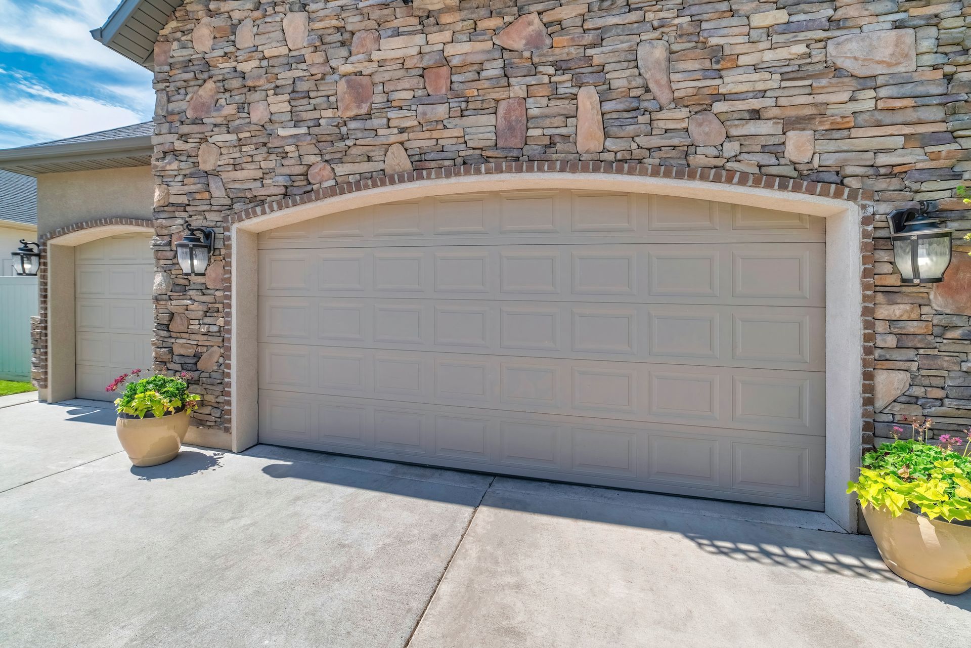 Stone-front house with beige garage door, potted plants, and bright driveway in sunlight