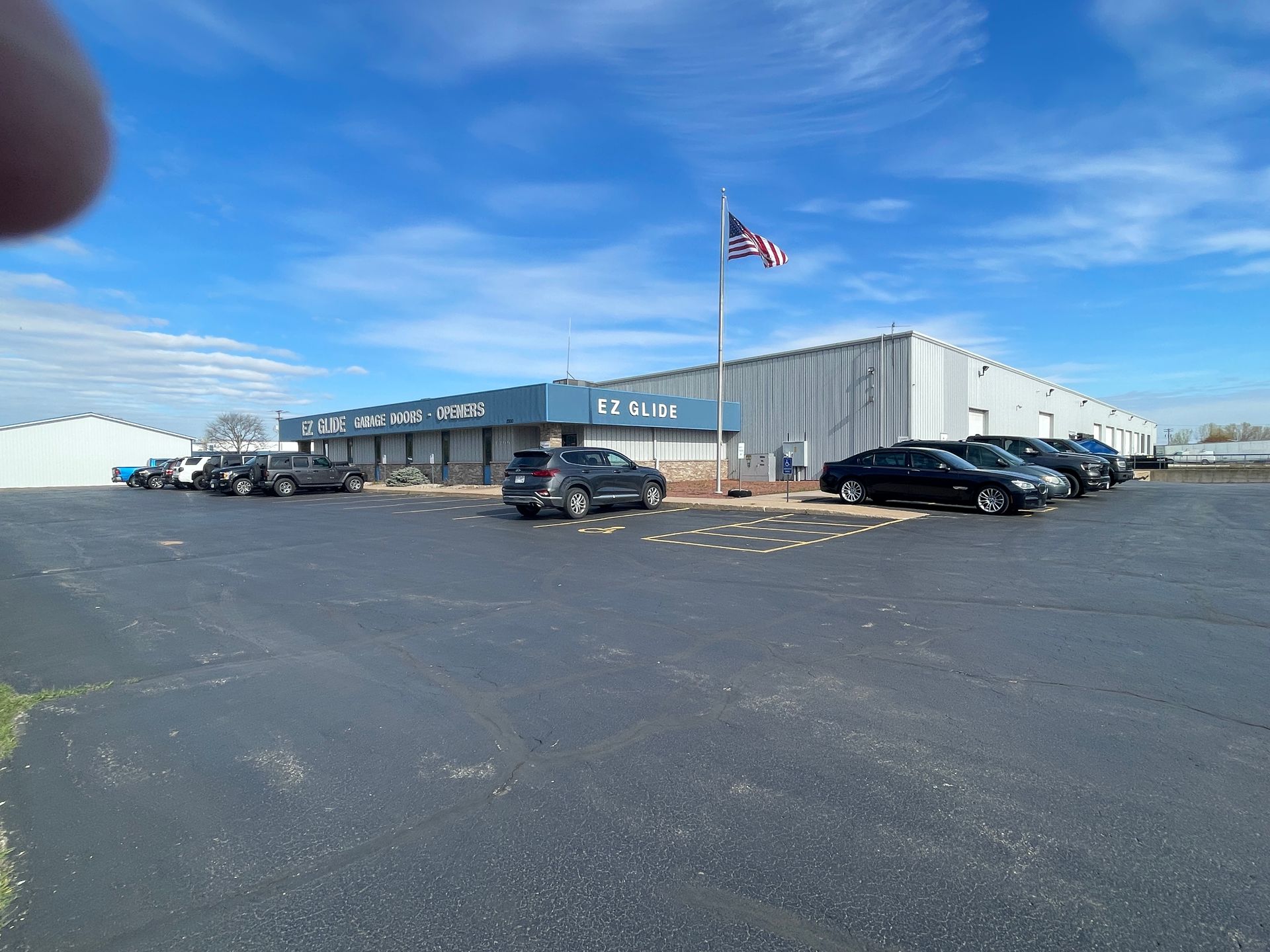 Wide parking lot in front of a gray industrial building with a U.S. flag and several parked cars under a blue sky