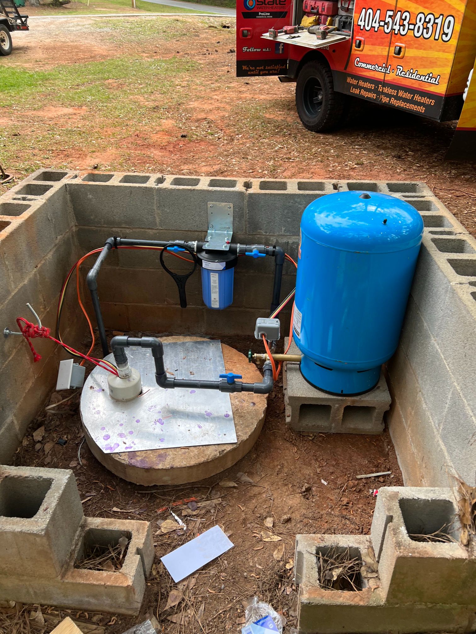 Well water system in a concrete enclosure; includes a blue tank, filter, and piping. A truck is in the background.