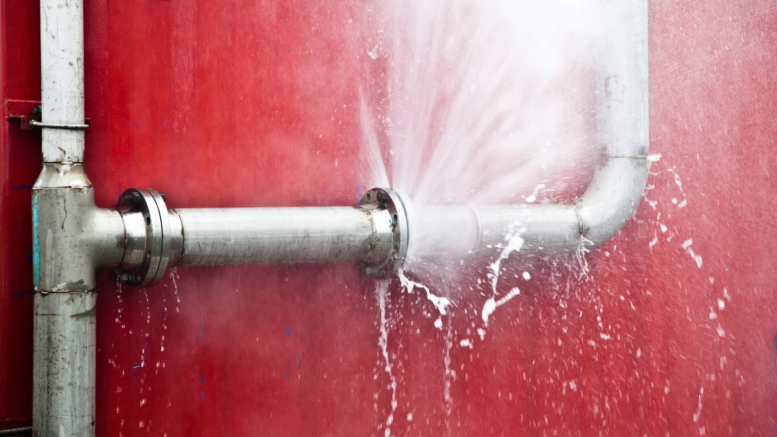 Leaking metal pipe on a red wall, spraying water across the surface
