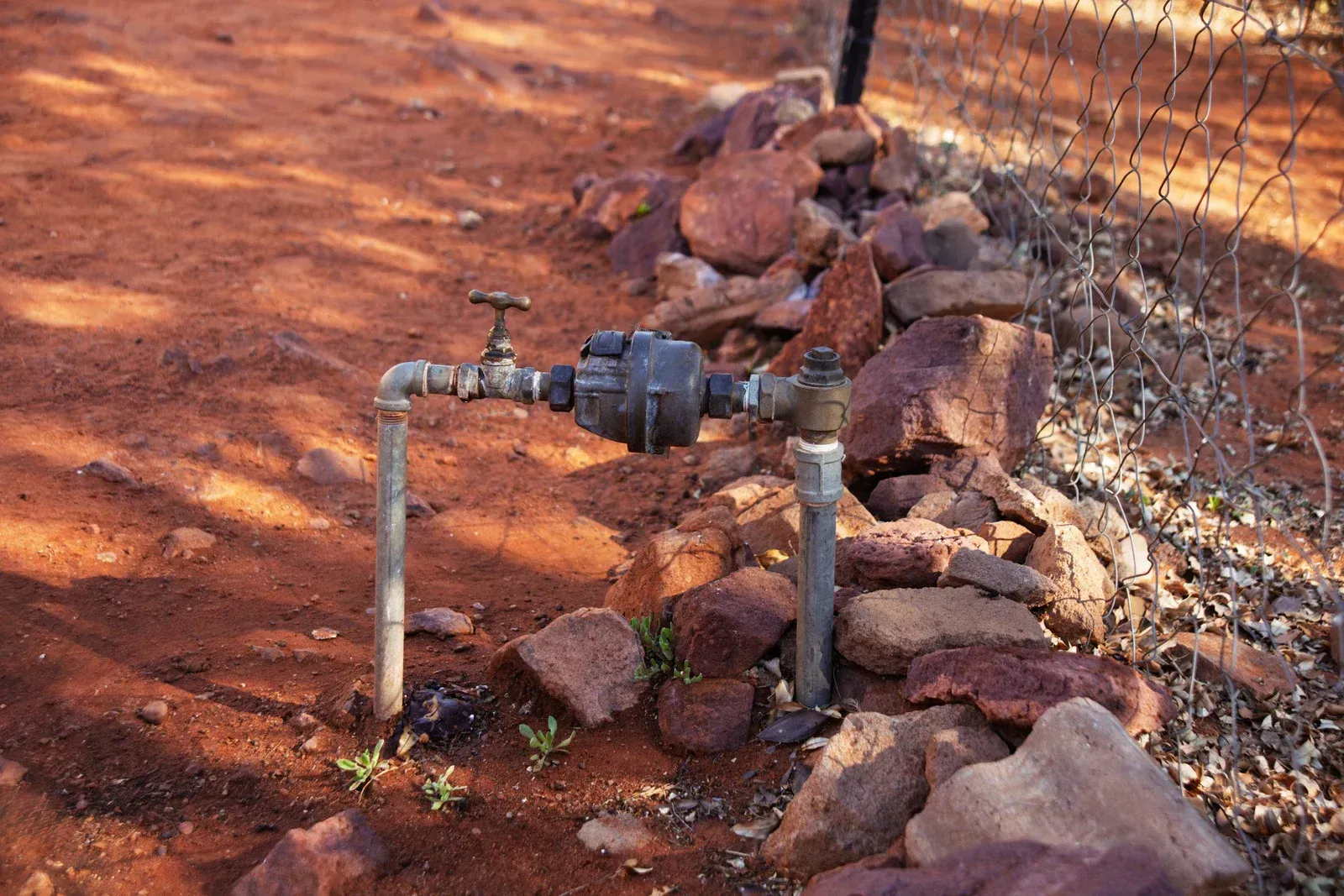 Water meter and pipes against a red dirt background, with rocks and a chain-link fence.