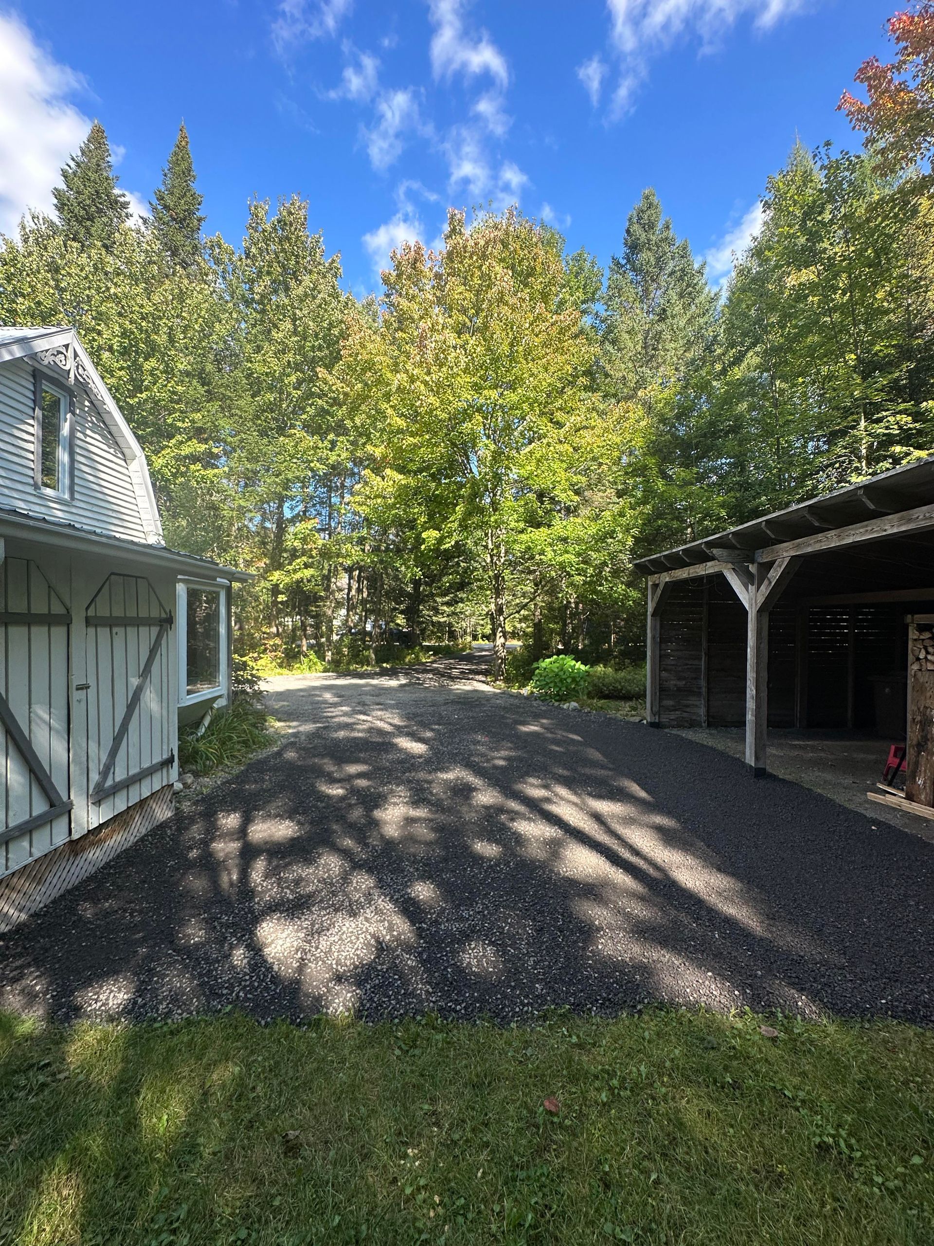 Une allée menant à un garage et une grange entourée d'arbres par une journée ensoleillée.