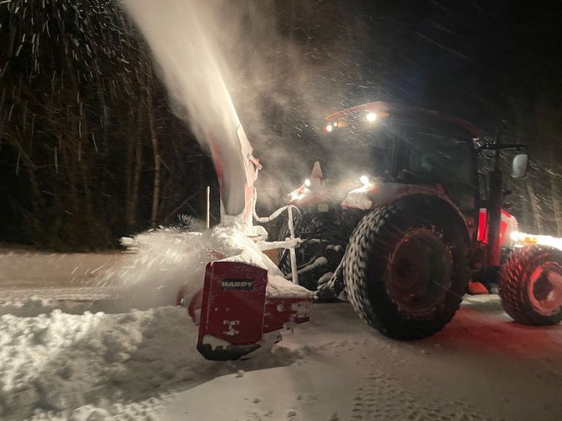 Un tracteur souffle de la neige la nuit sur une route enneigée.