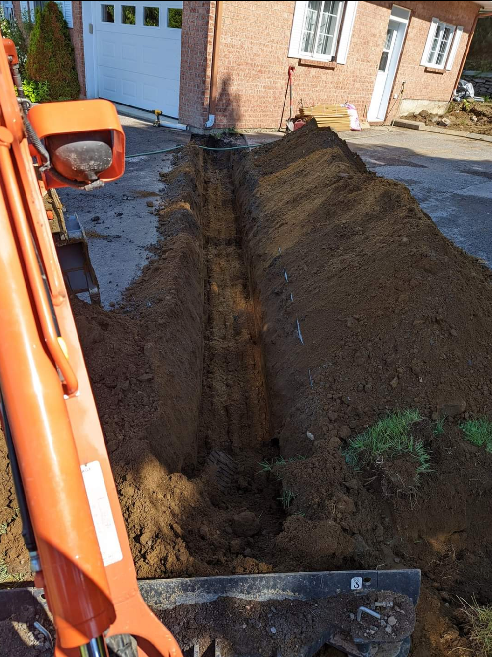 Une excavatrice orange creuse une tranchée devant une maison en brique.