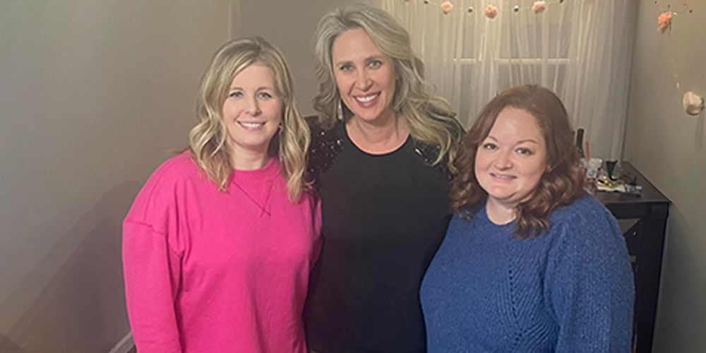 Three smiling women pose for a photo in a room with a decorated backdrop. One in pink, one in black, and one in blue.
