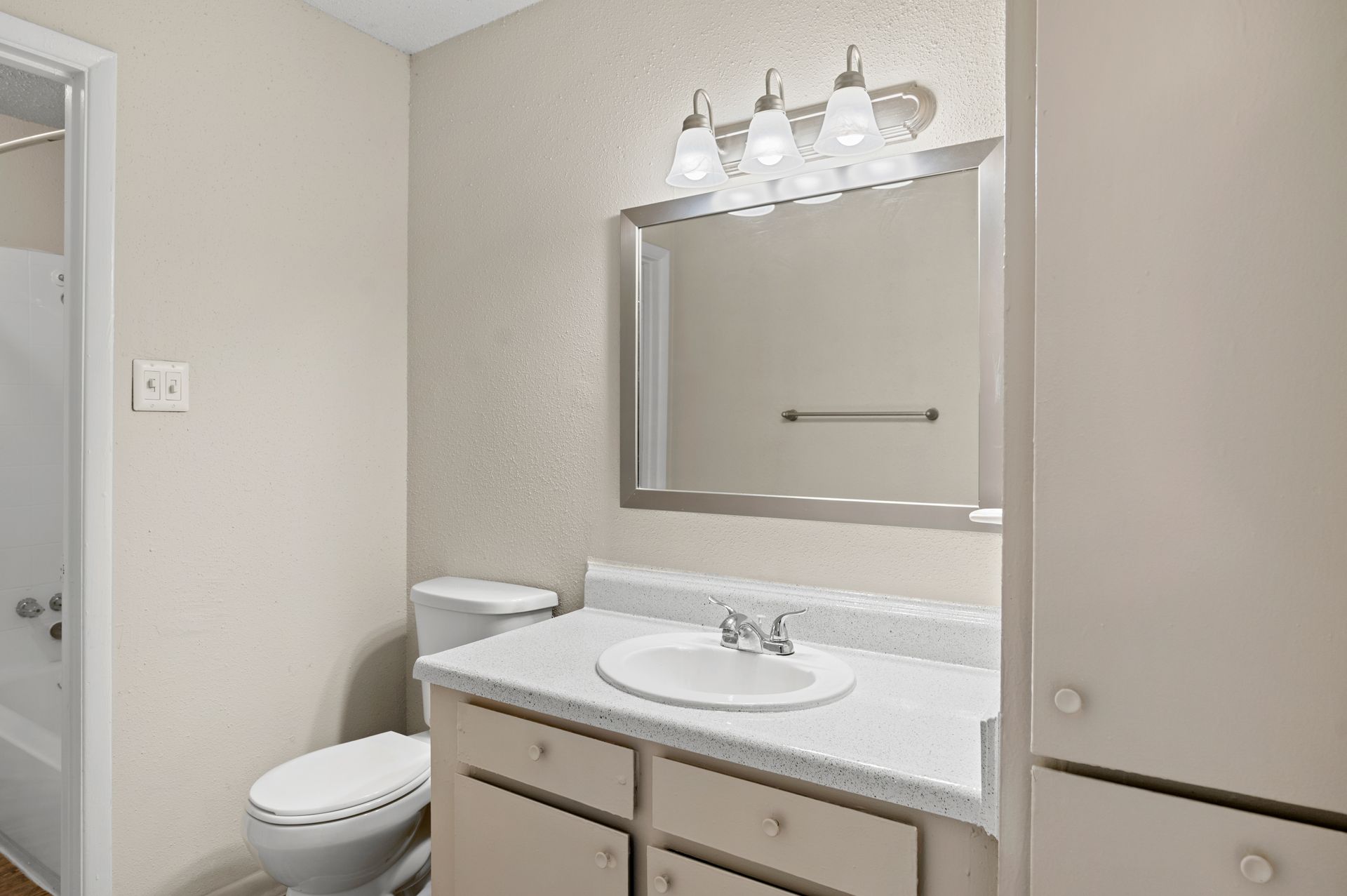 Bathroom with a toilet, sink, mirror, and cabinet. Neutral colors, overhead lighting, and a bathtub visible.