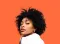 Young Black woman with curly hair against a bright orange background; looking towards the camera.