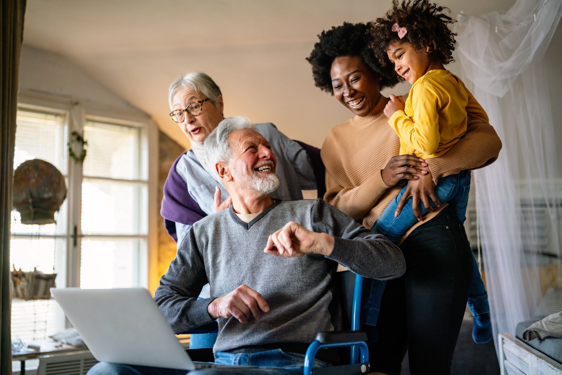 Family gathered around a man in a wheelchair looking at a laptop, smiling. Woman holding a child, other woman in background.