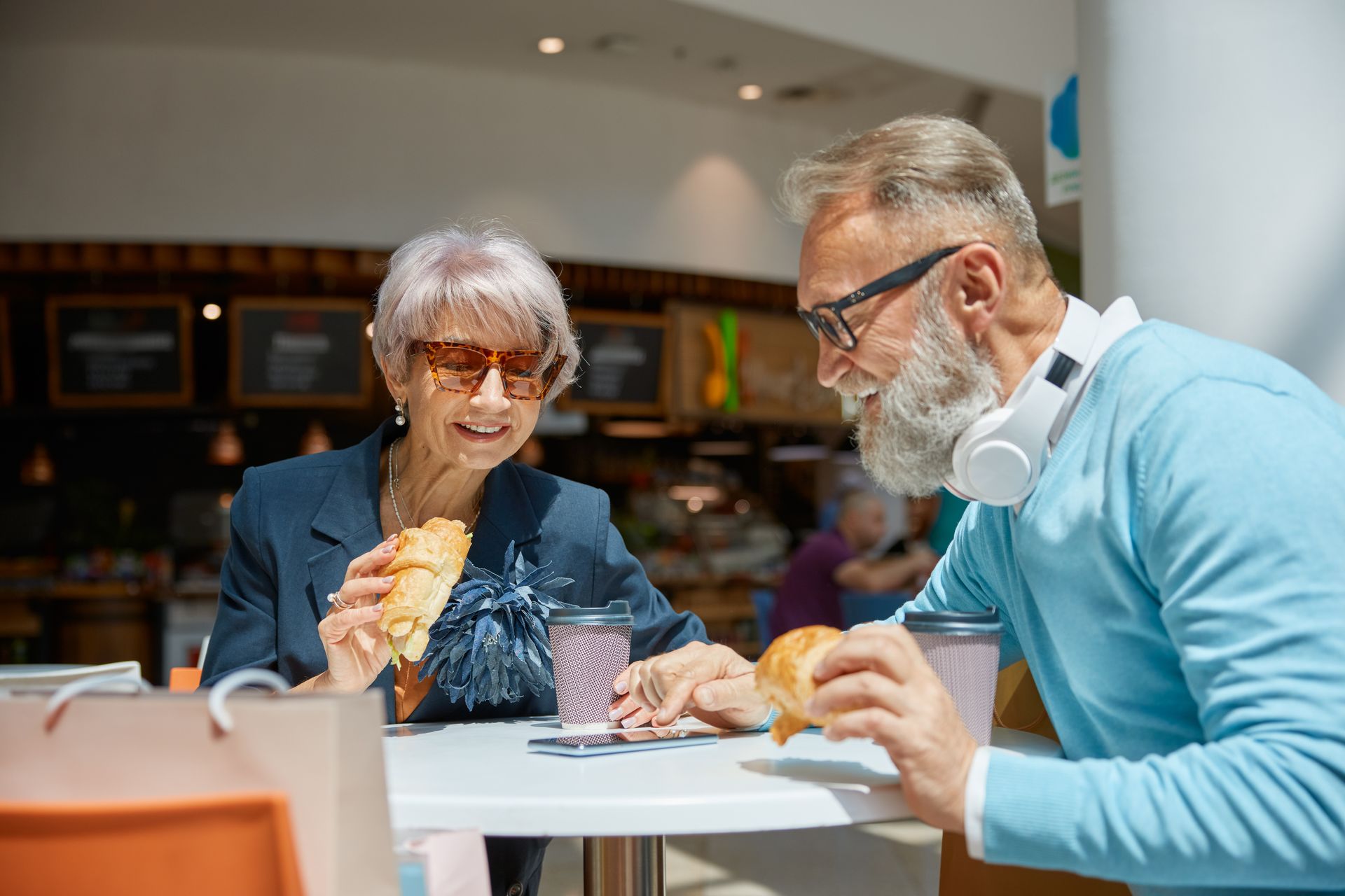 An older couple eating, looking at a phone, and drinking coffee at a table in a cafe.