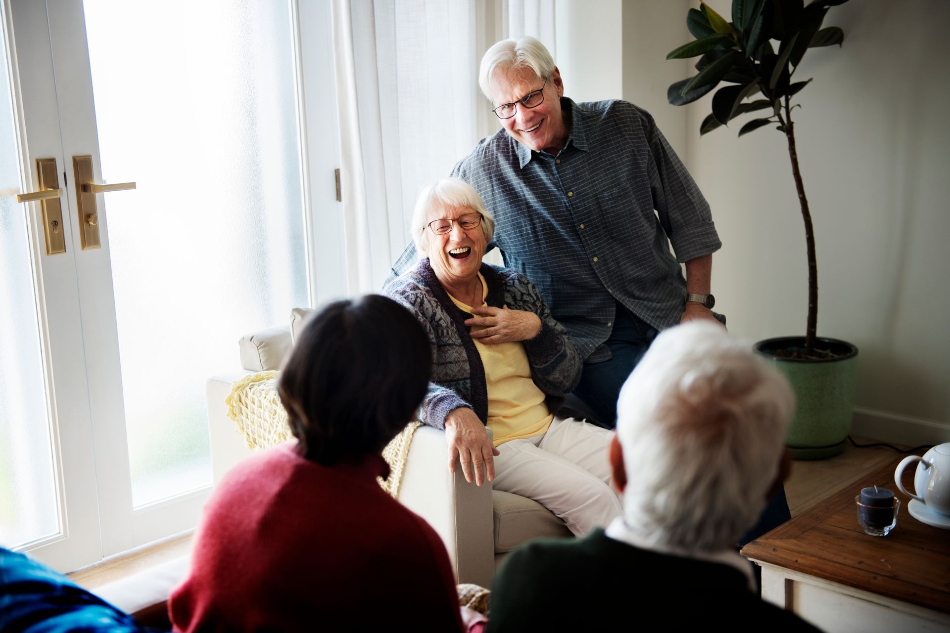 Group of people in a brightly lit living room; senior woman laughs while others converse and smile.