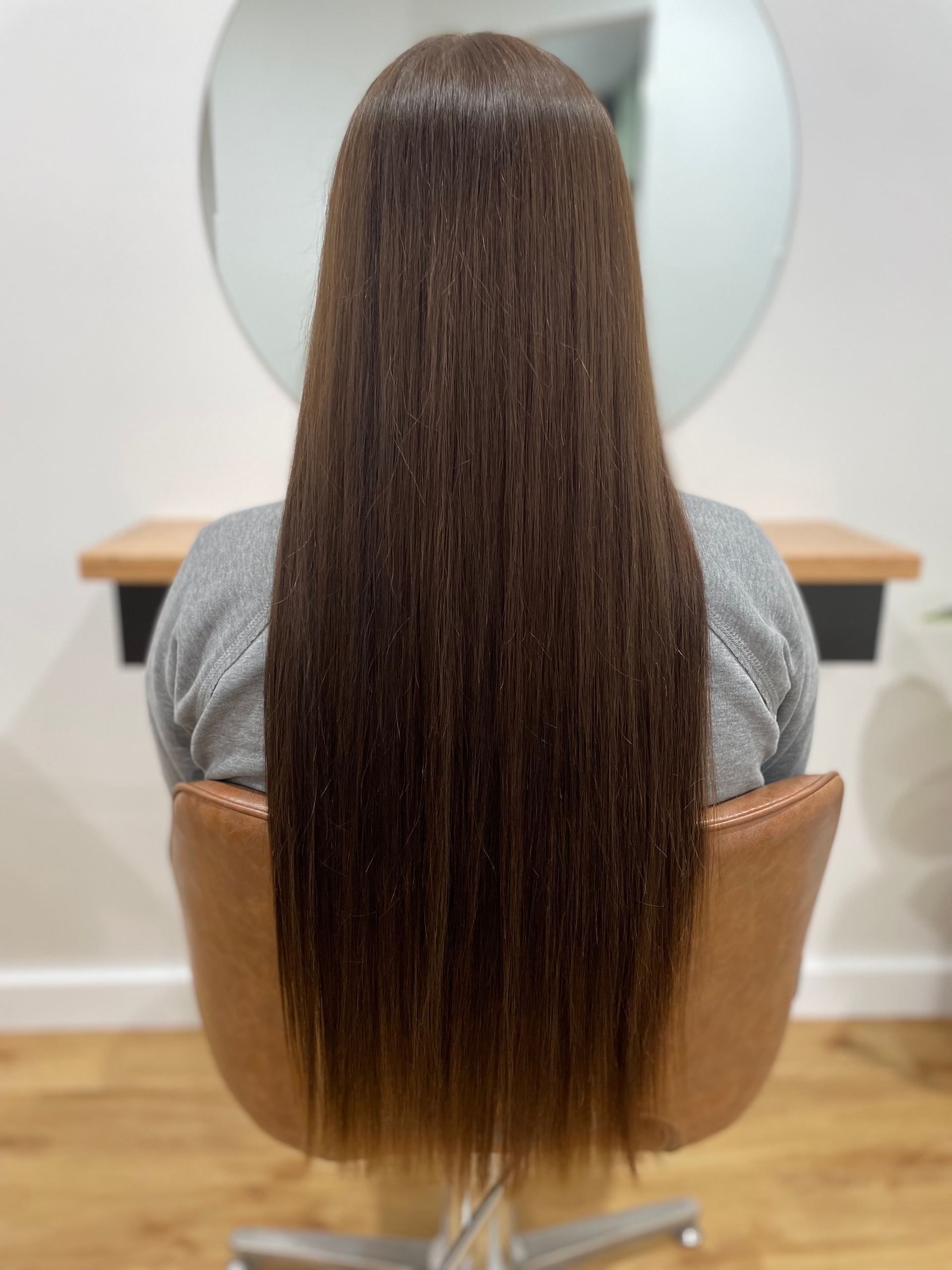 Person seated in a salon chair, facing away, with long, straight, brown hair, against a neutral background — Clippers Hair Salon in Taree, NSW