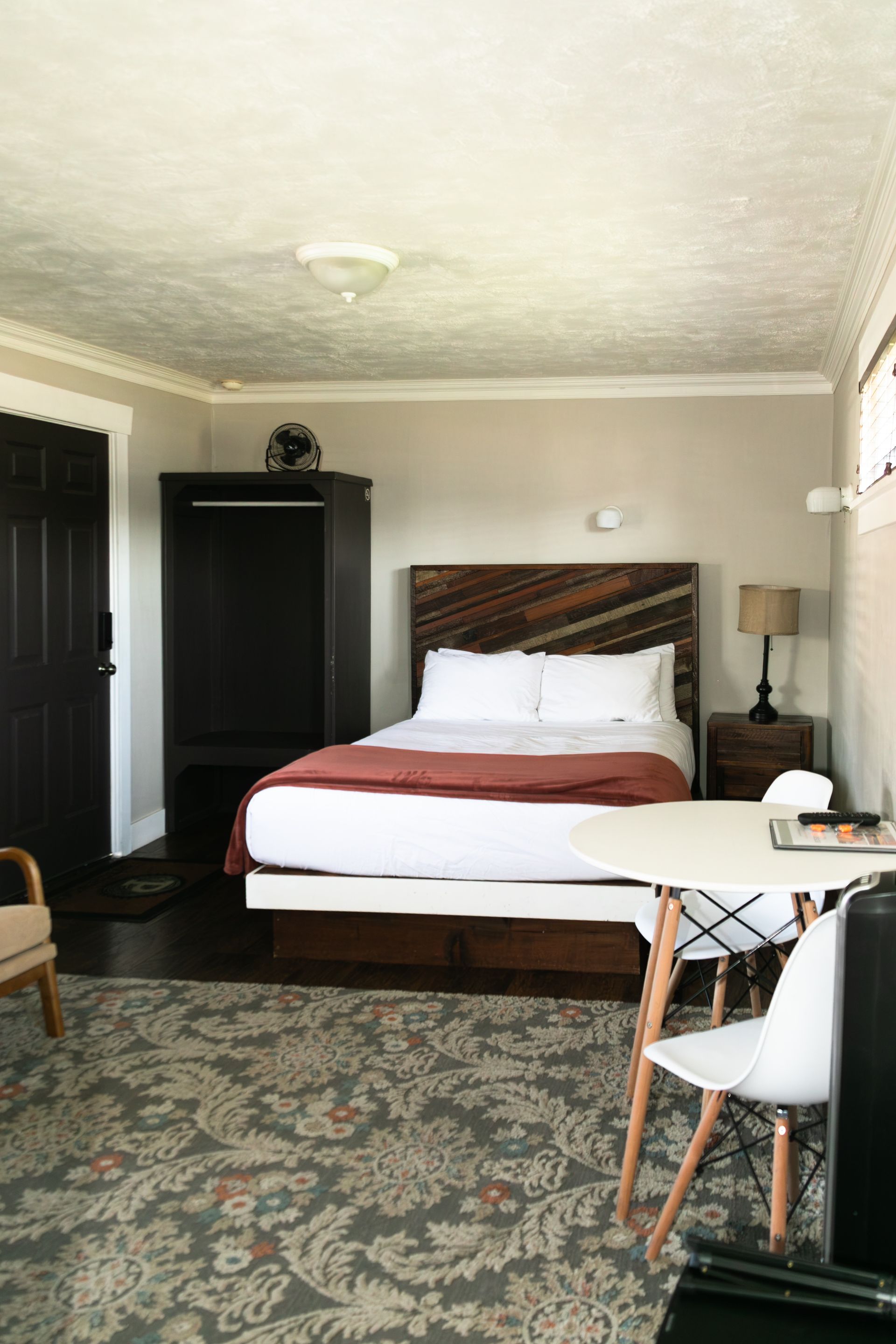 Cozy bedroom with a bed, patterned rug, dark armoire, and small table by the window.