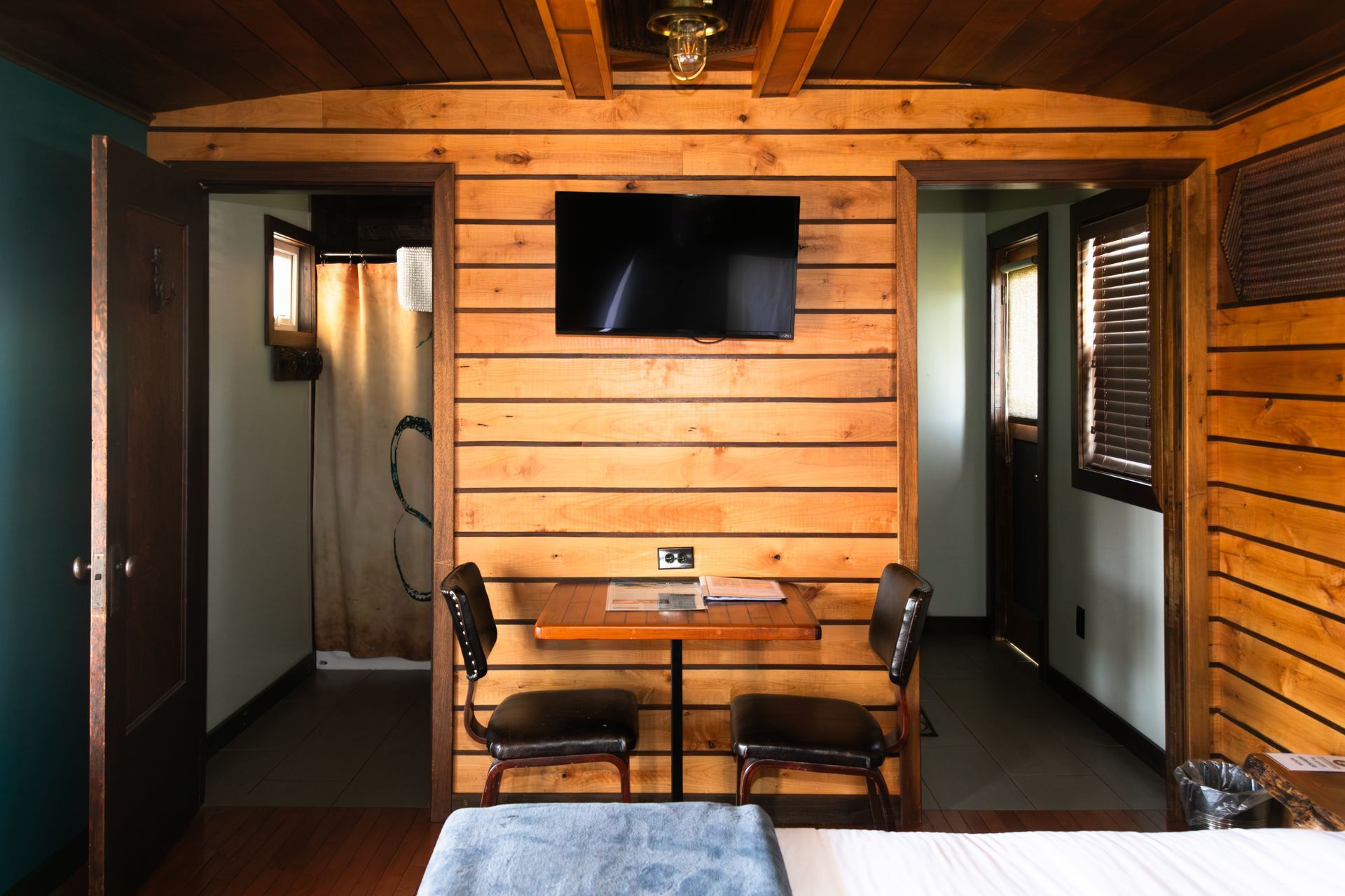 Cozy wood-paneled room with wall TV, two chairs and a small table, hallway doors on both sides.