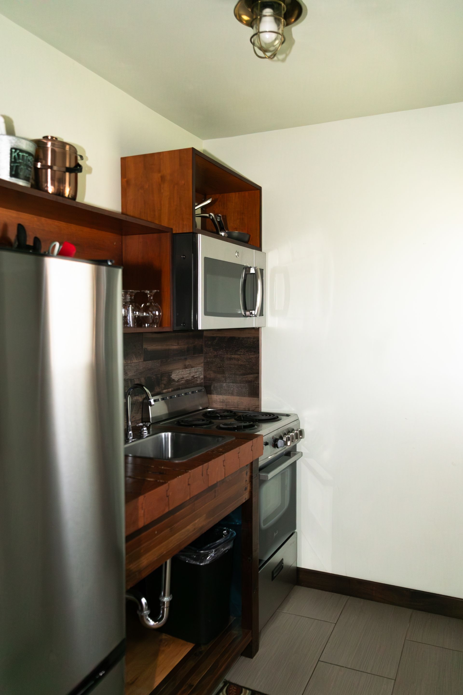 Small kitchen with stainless steel fridge, wooden cabinets, stove, and sink against white walls.
