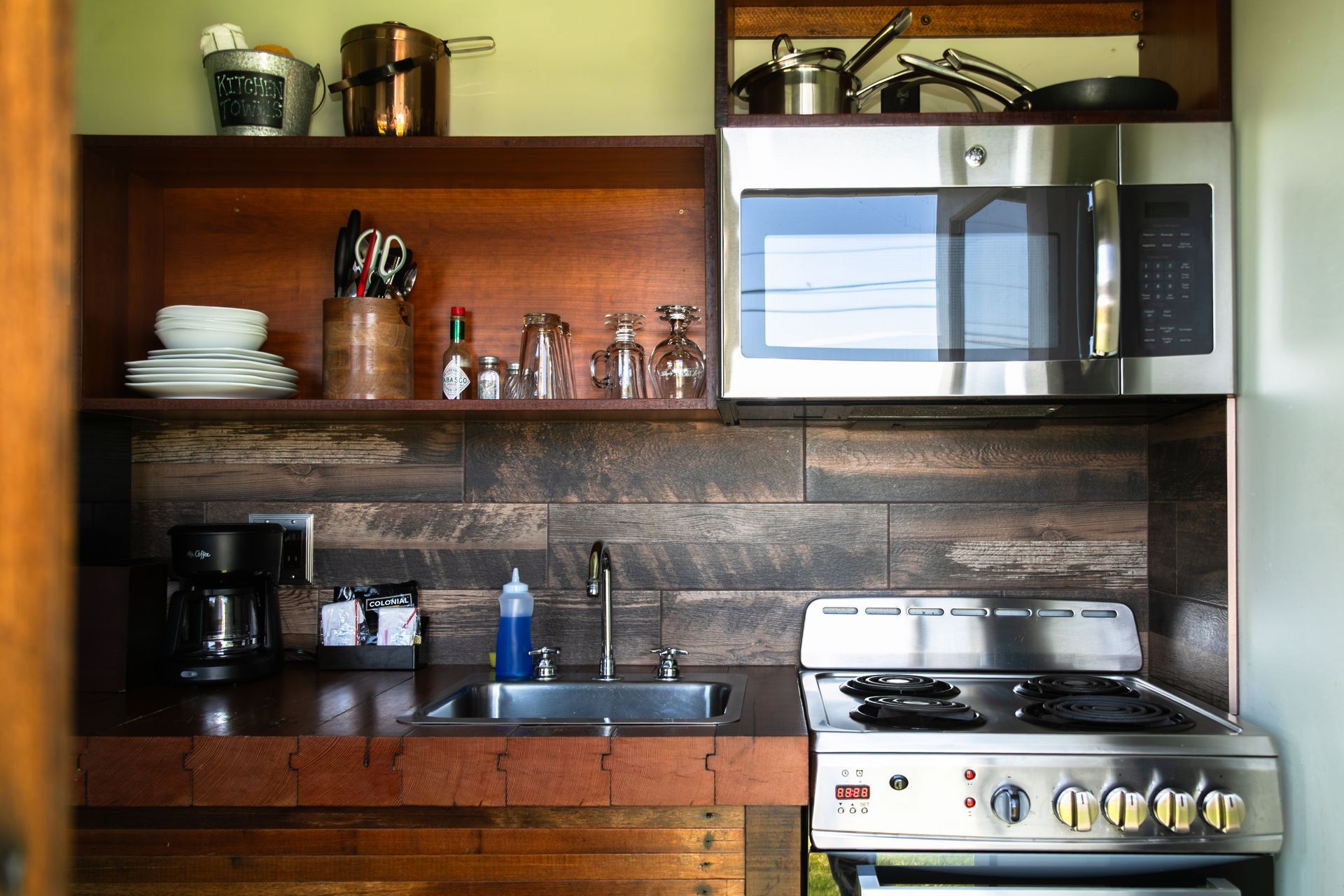 Small kitchen with wooden shelves, dishes, sink, coffee maker, microwave, and gas stove.
