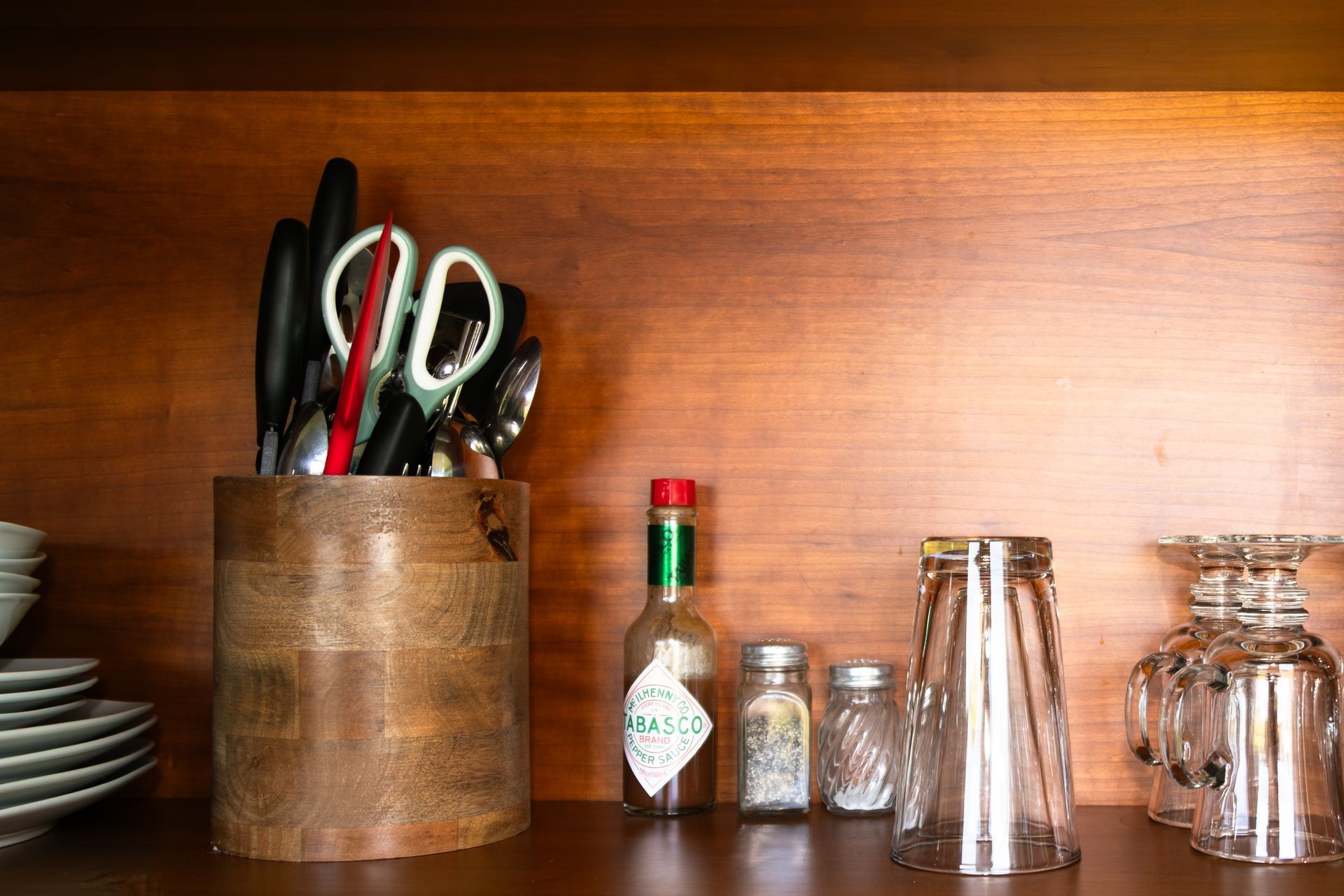 Kitchen counter with utensils in a wooden holder, spice bottles, and glasses on a wooden shelf