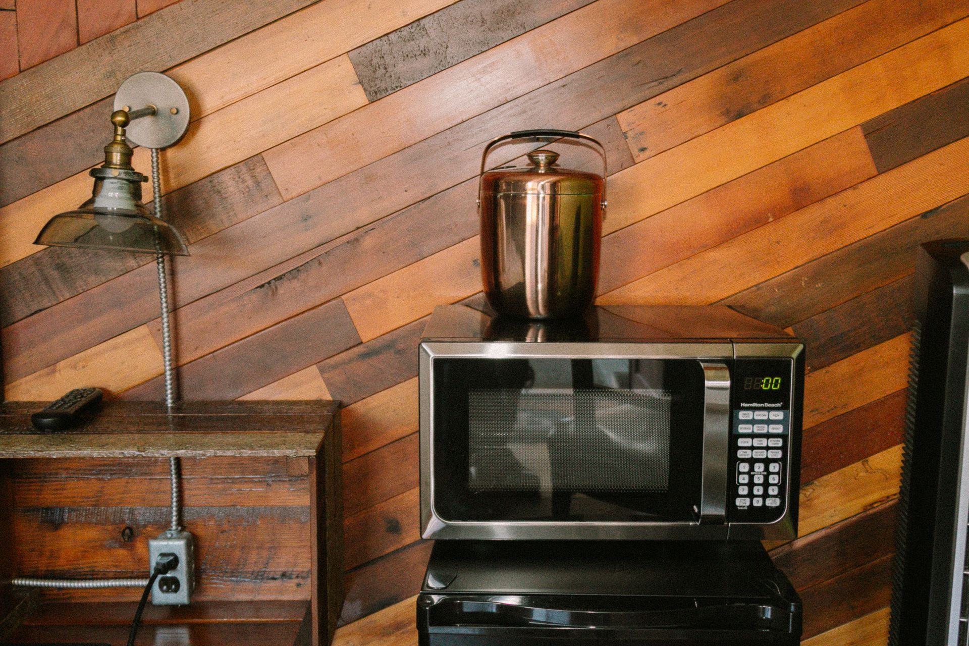 Microwave on a counter with a metal blender, beside a wooden cabinet and wall lamp in a rustic kitchen area