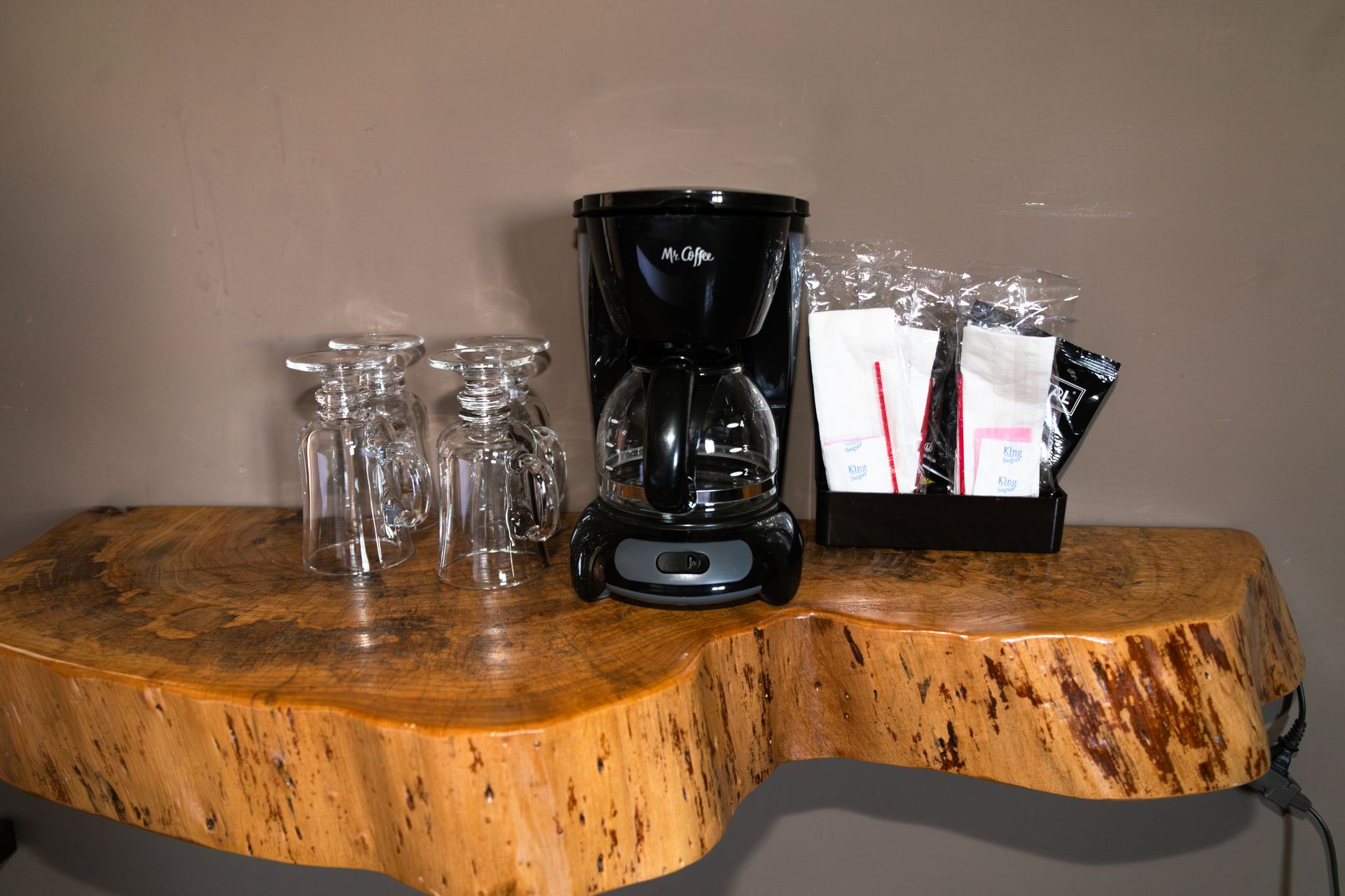 Black coffee maker beside stacked glass cups and sugar packets on a rustic wood table