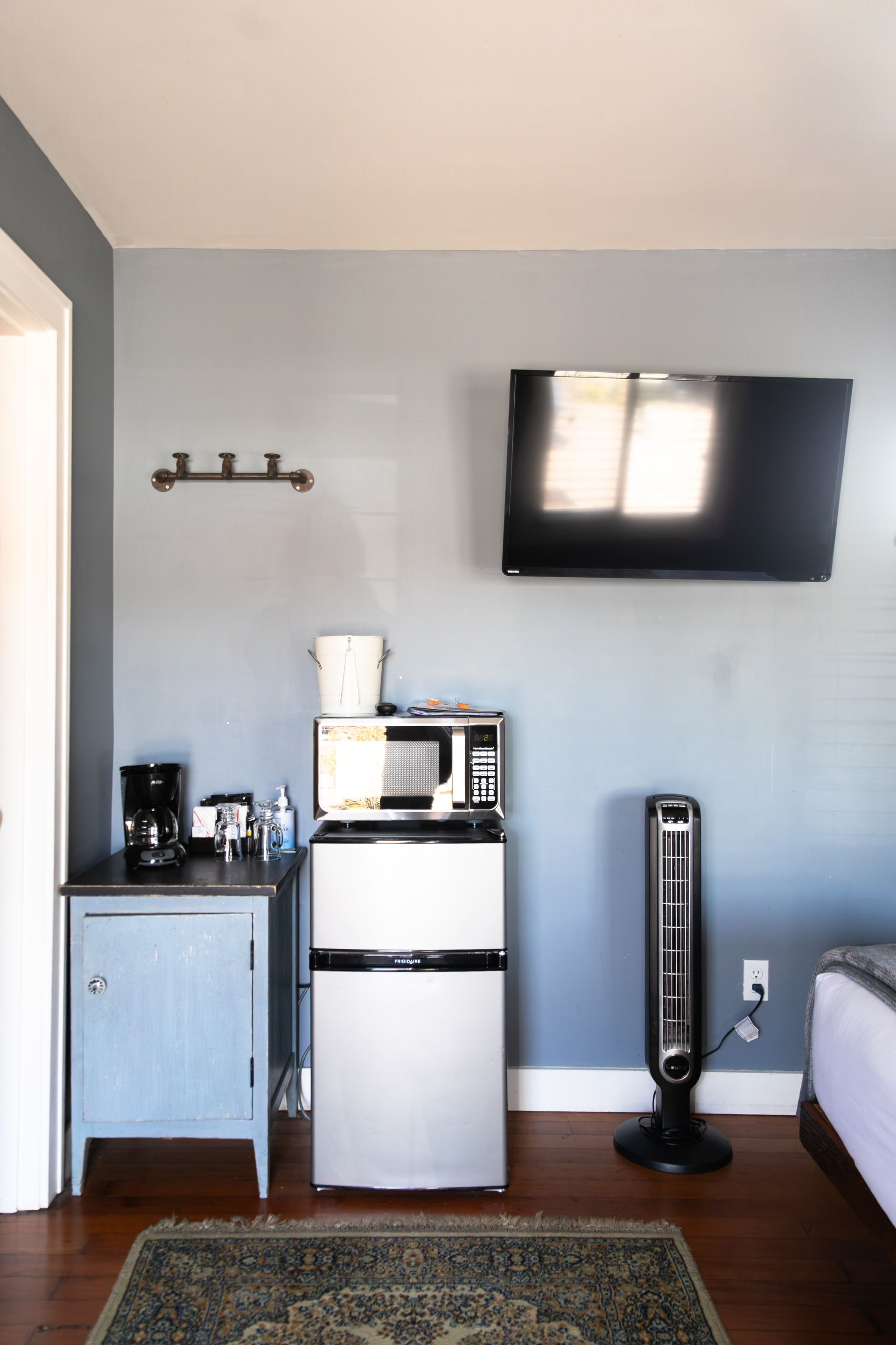 Small room with blue-gray walls, wall-mounted TV, mini fridge, coffee maker, cabinet, and tower fan on wood floor.