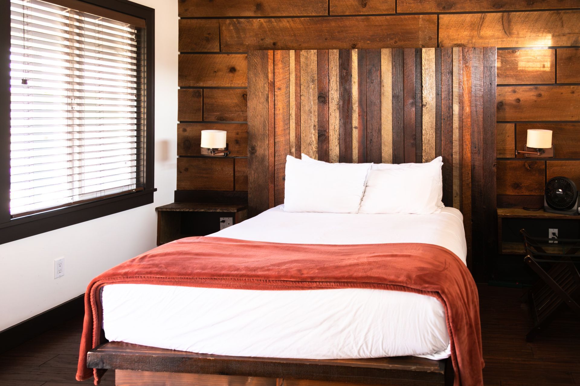 Wood-paneled bedroom with a bed, orange blanket, bedside lamps, and a window with blinds