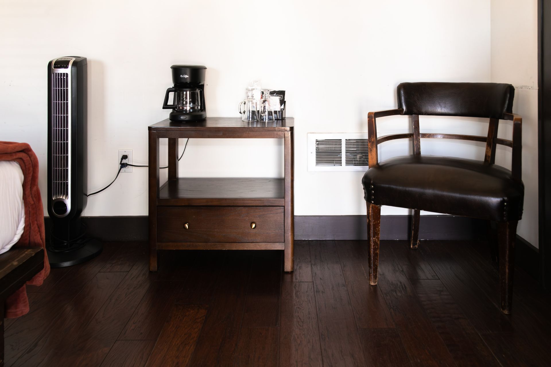 Modern room with a black chair, small wooden table, coffee maker, and tall fan on dark hardwood floor