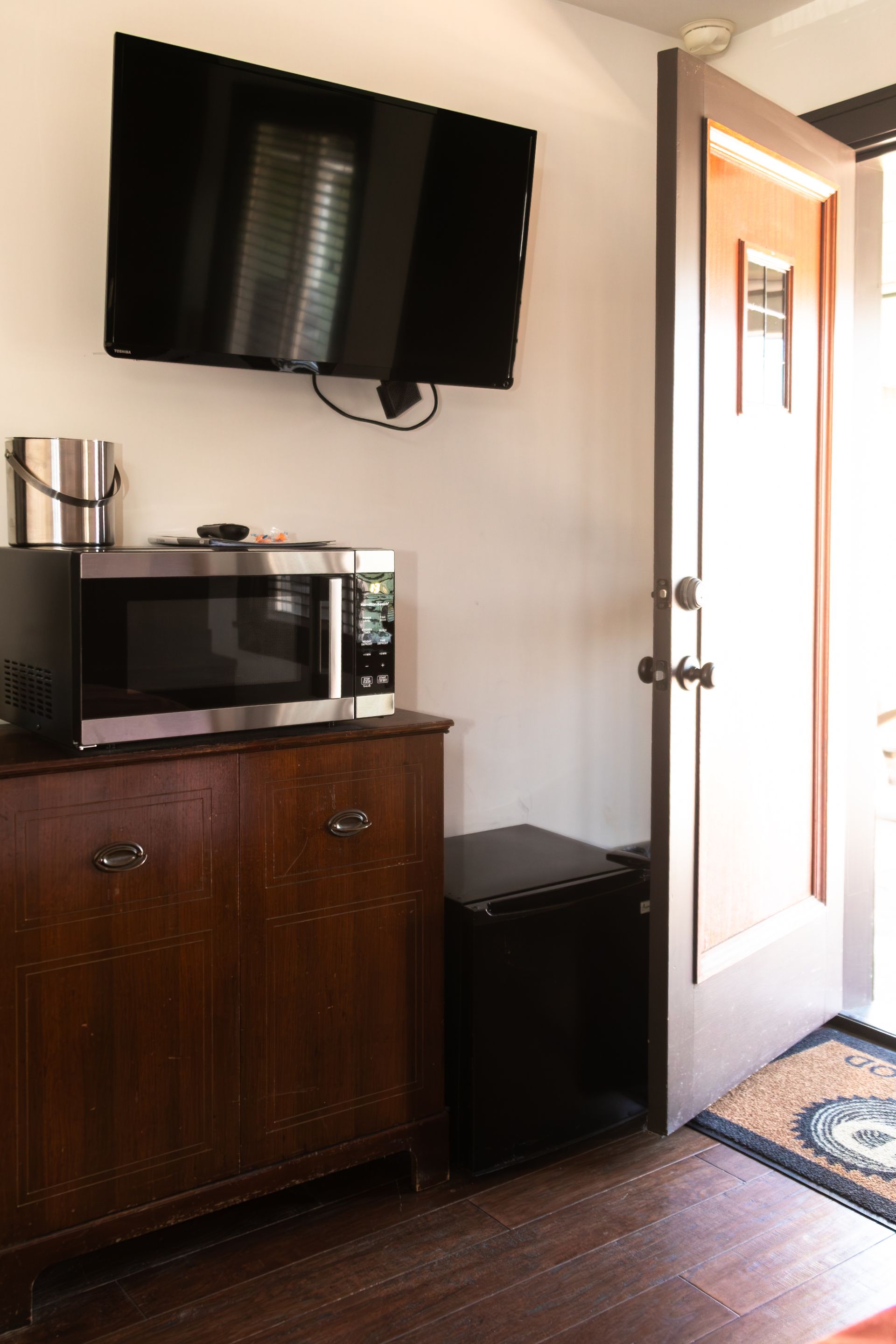 Small room with wall-mounted TV, microwave on cabinet, mini fridge, and open door to bright hallway.
