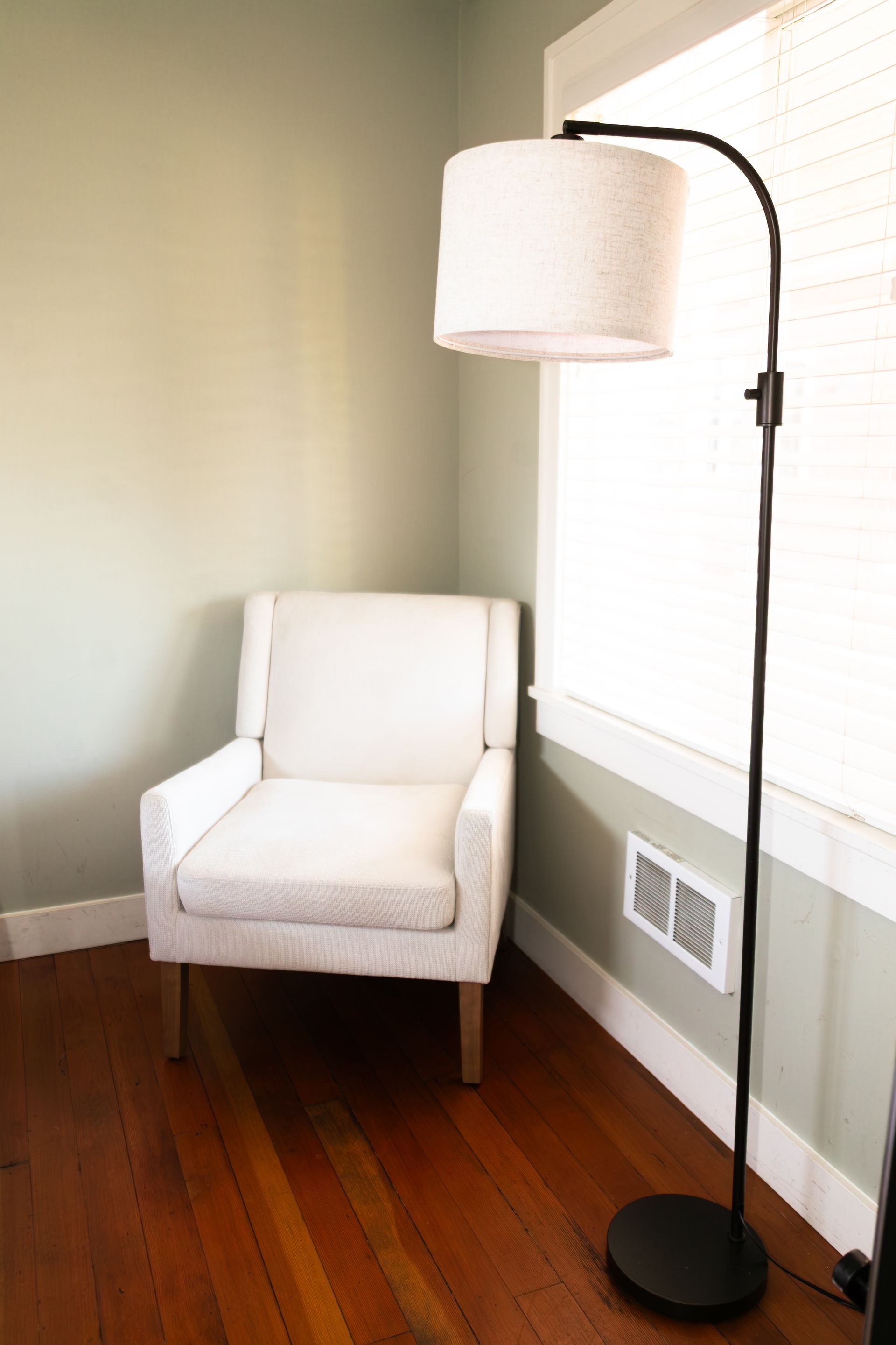 White armchair beside a black floor lamp in a bright corner with wood floor and window blinds