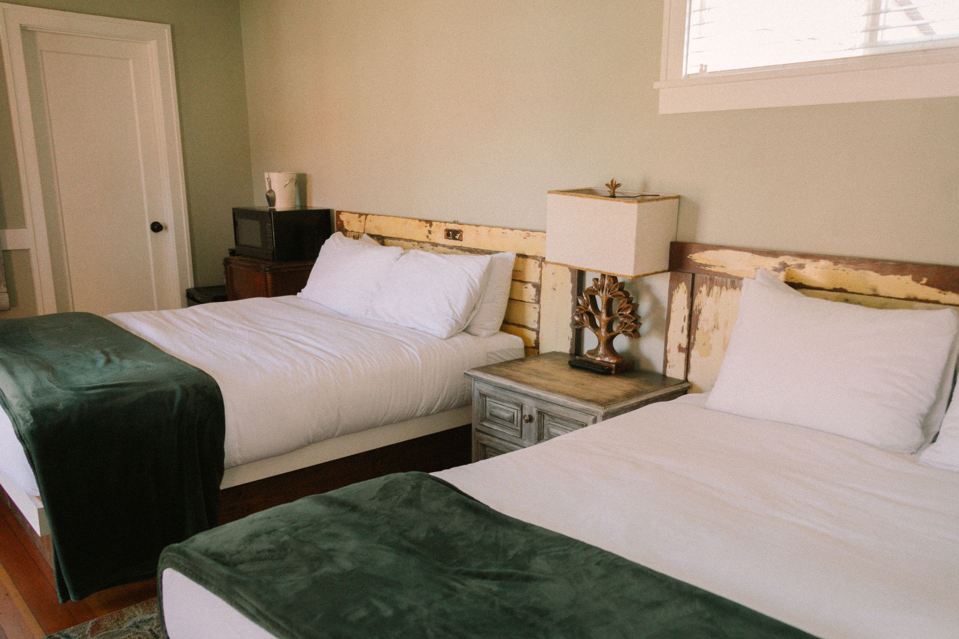 Two twin beds with green blankets in a beige hotel room, separated by a nightstand and lamp.