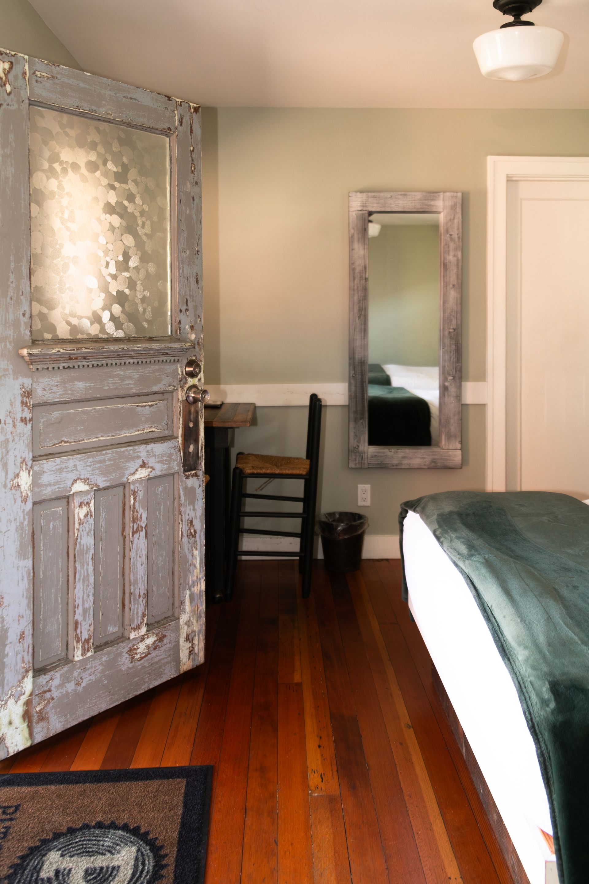 Bedroom with distressed gray door, mirror, chair, wood floor, and bed with green blanket