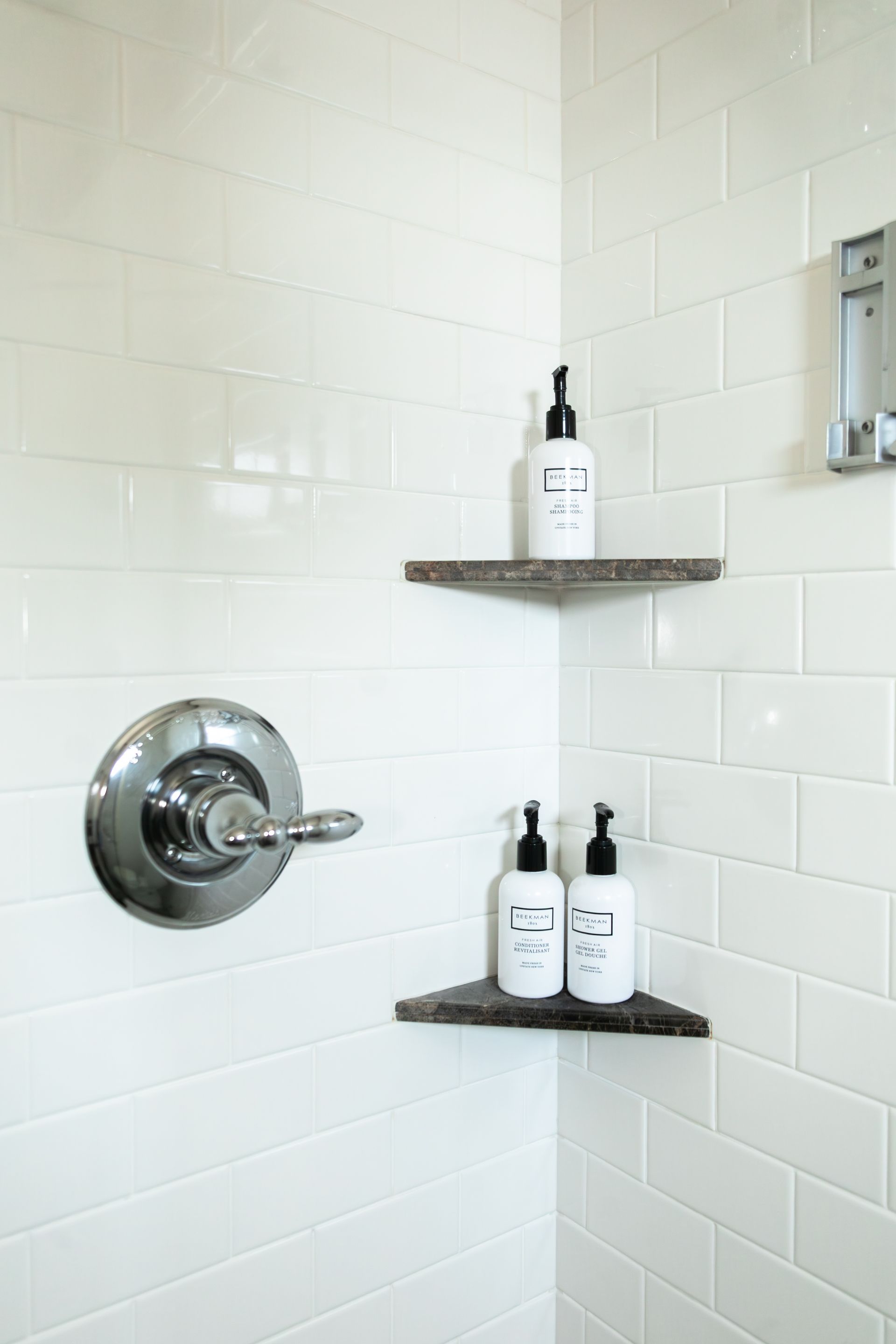 White tiled shower corner with chrome control and two corner shelves holding soap bottles.