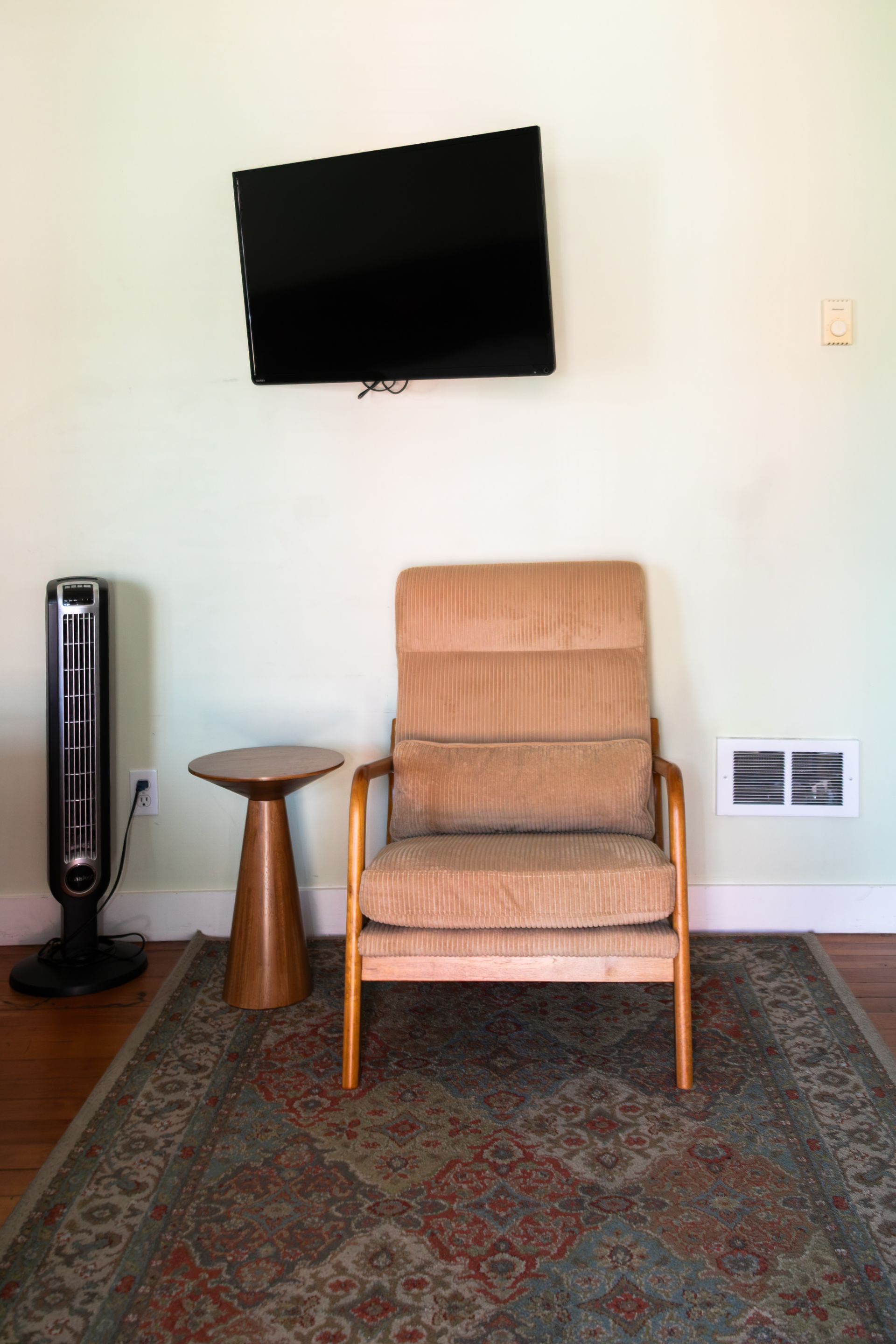 Living room with wall-mounted TV, tan armchair, rug, and tower fan on a white wall.