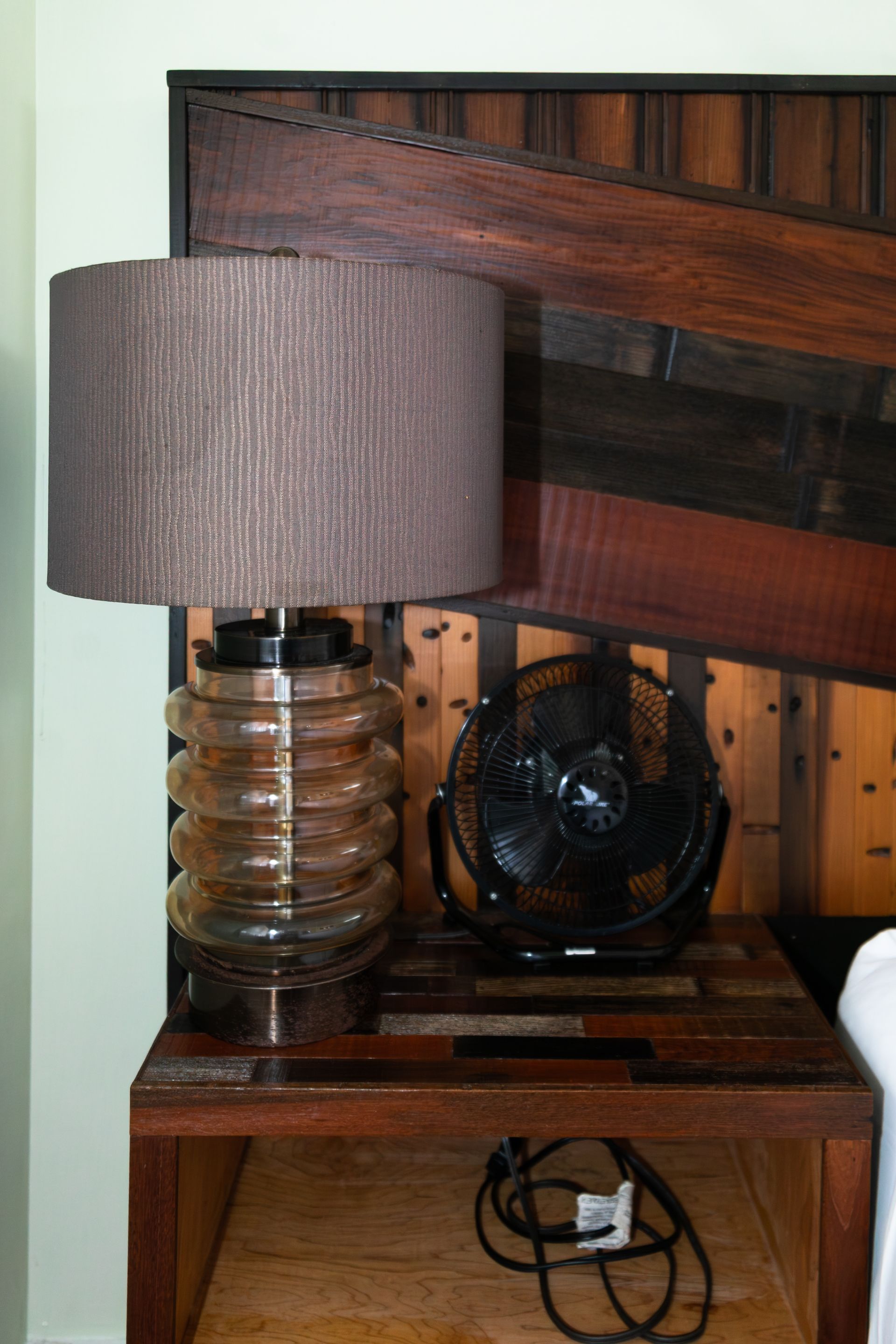 Wooden bedside table with gray lamp, black fan, and coiled cord beneath a dark wood headboard