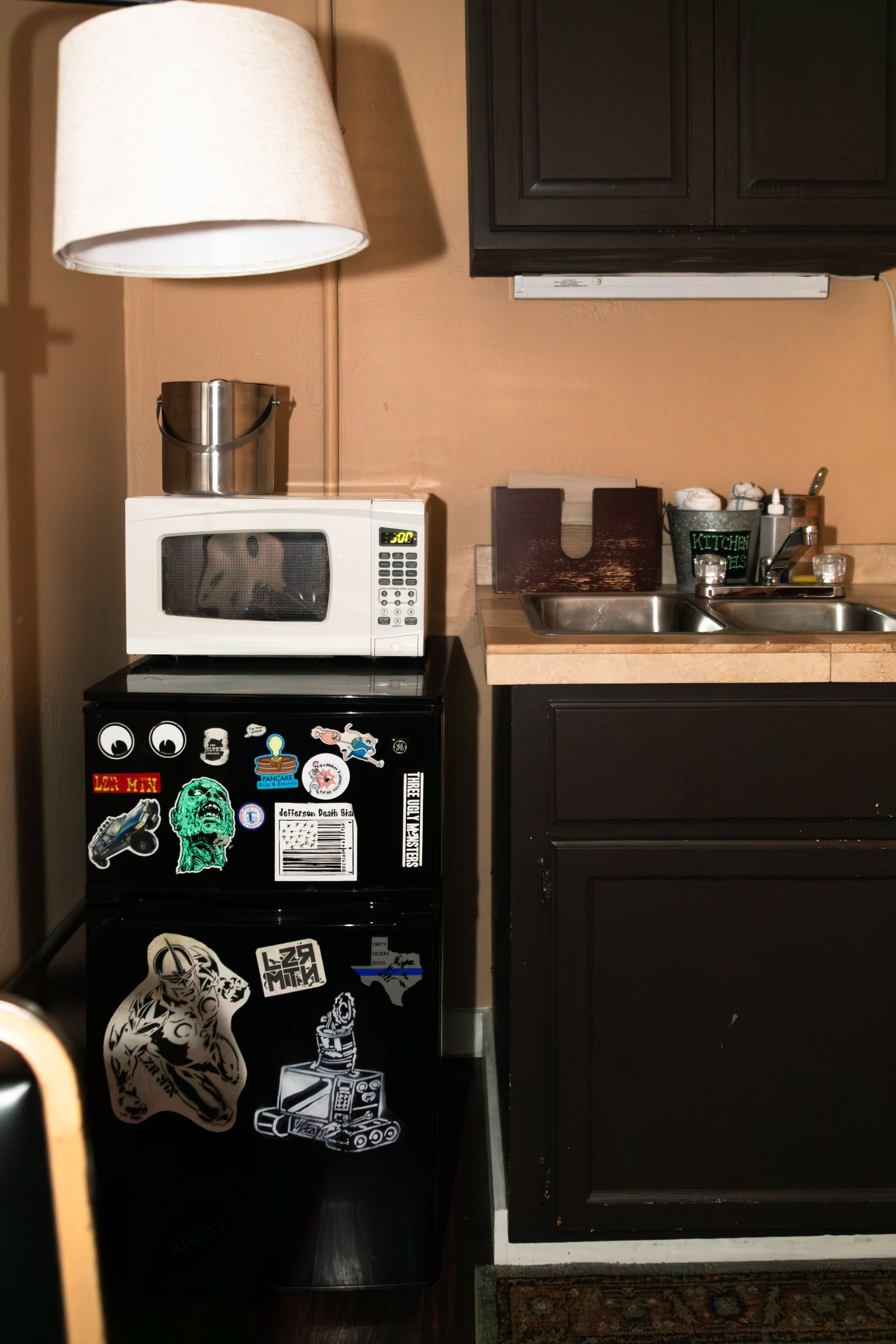 Small kitchen with black fridge covered in magnets, microwave, cabinets, and a lamp overhead.