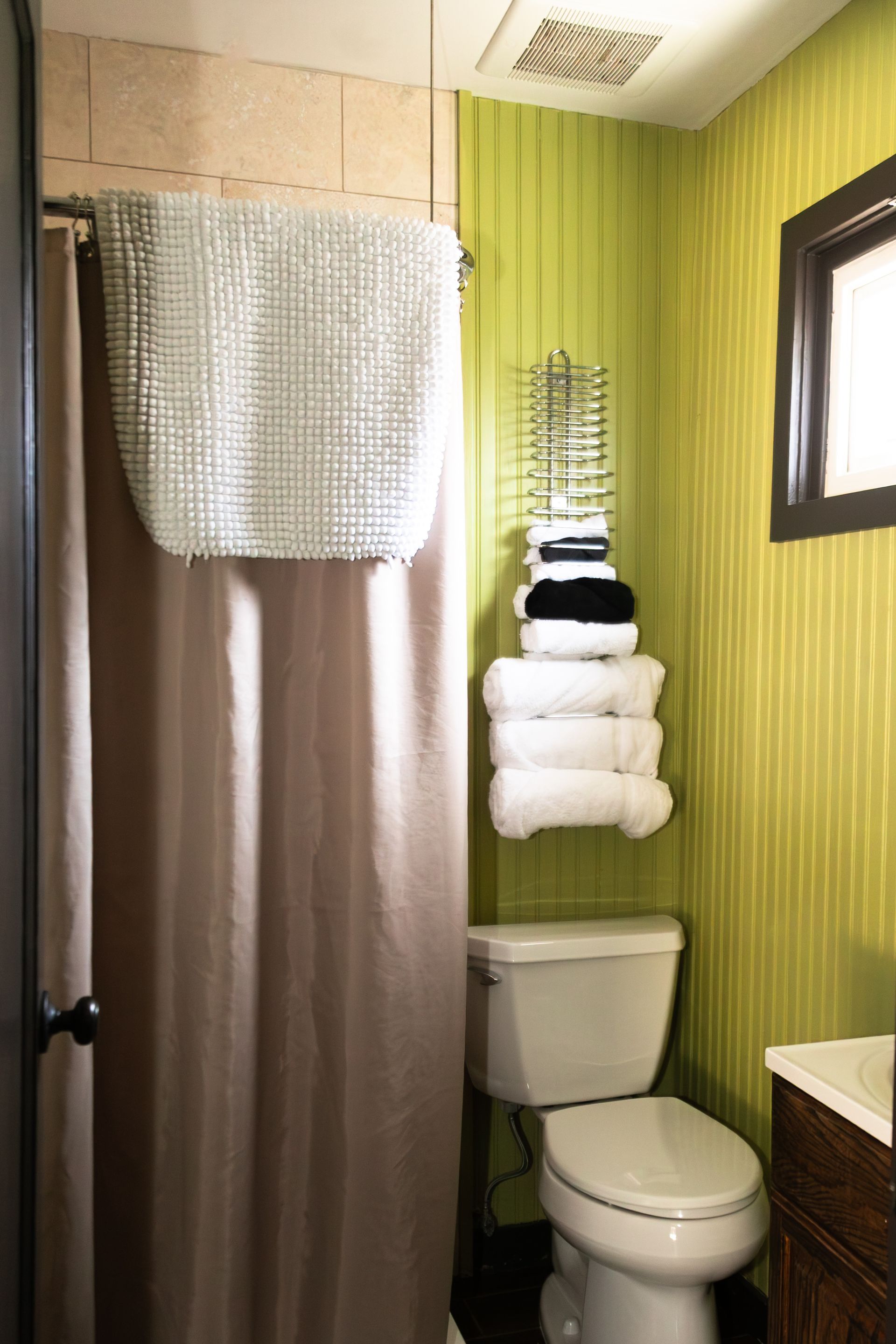 Small bathroom with shower curtain, toilet, stacked towels, green striped walls, and a window.