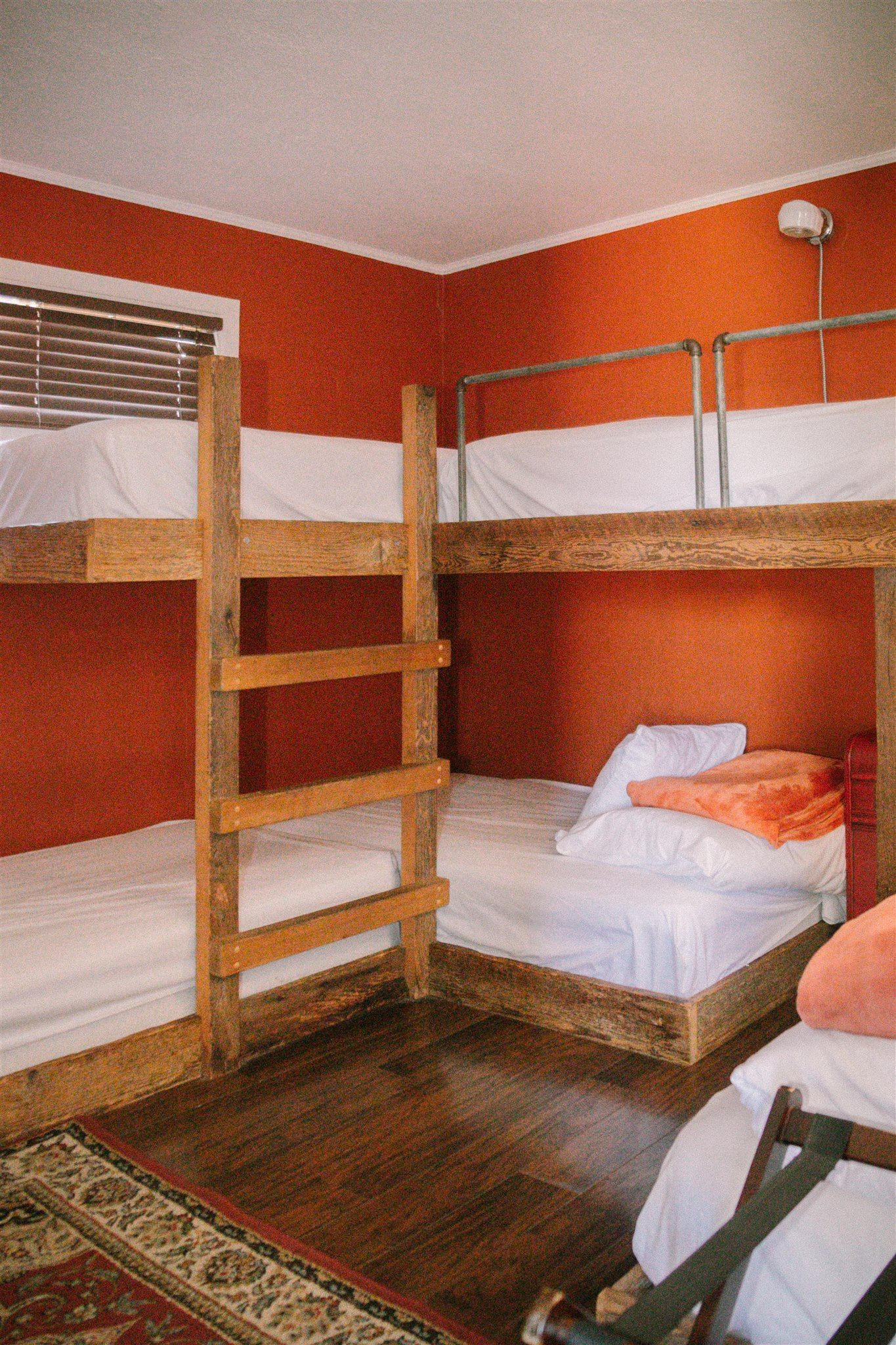 Wooden bunk bed in a small orange-painted bedroom with a patterned rug and window blinds