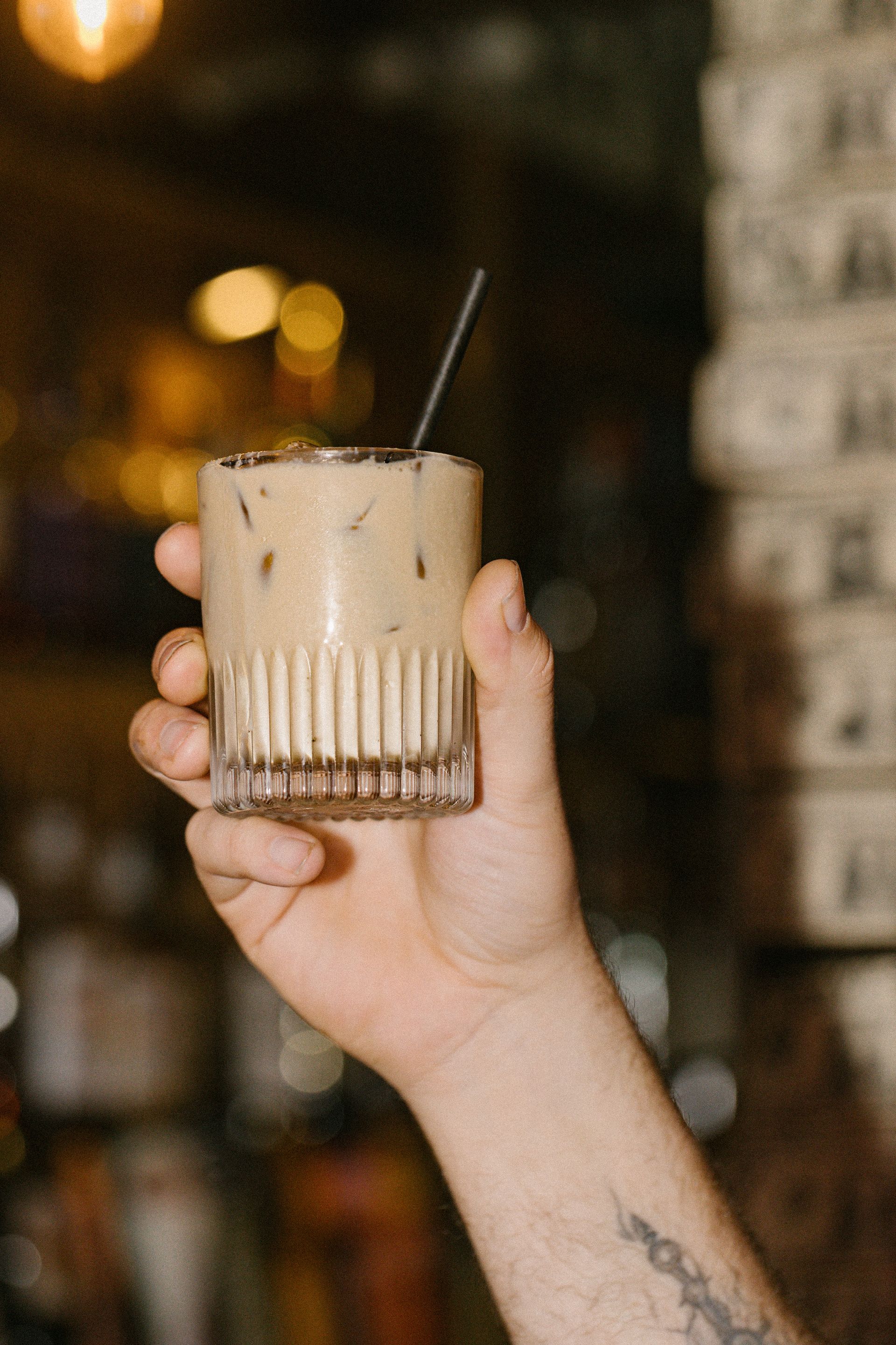 Hand holding a tan iced drink in a ribbed glass with a straw, against a blurred warm café background
