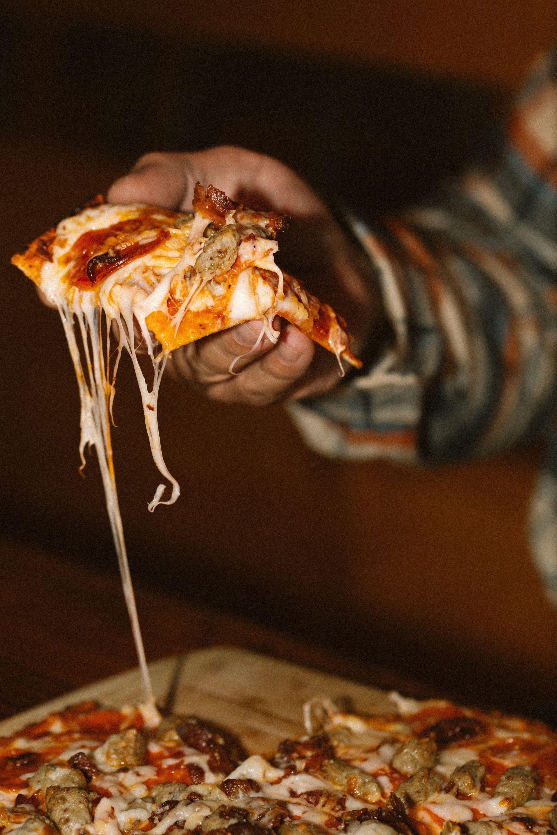Hand holding a slice of cheesy pizza above a pizza box, with melted cheese stretching down