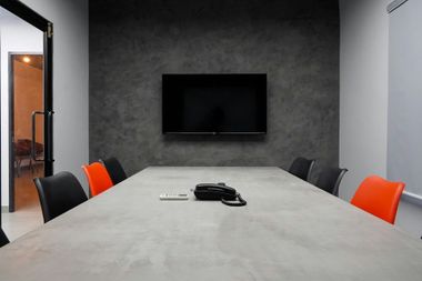 Conference room with gray table, black TV on gray wall, black and orange chairs.