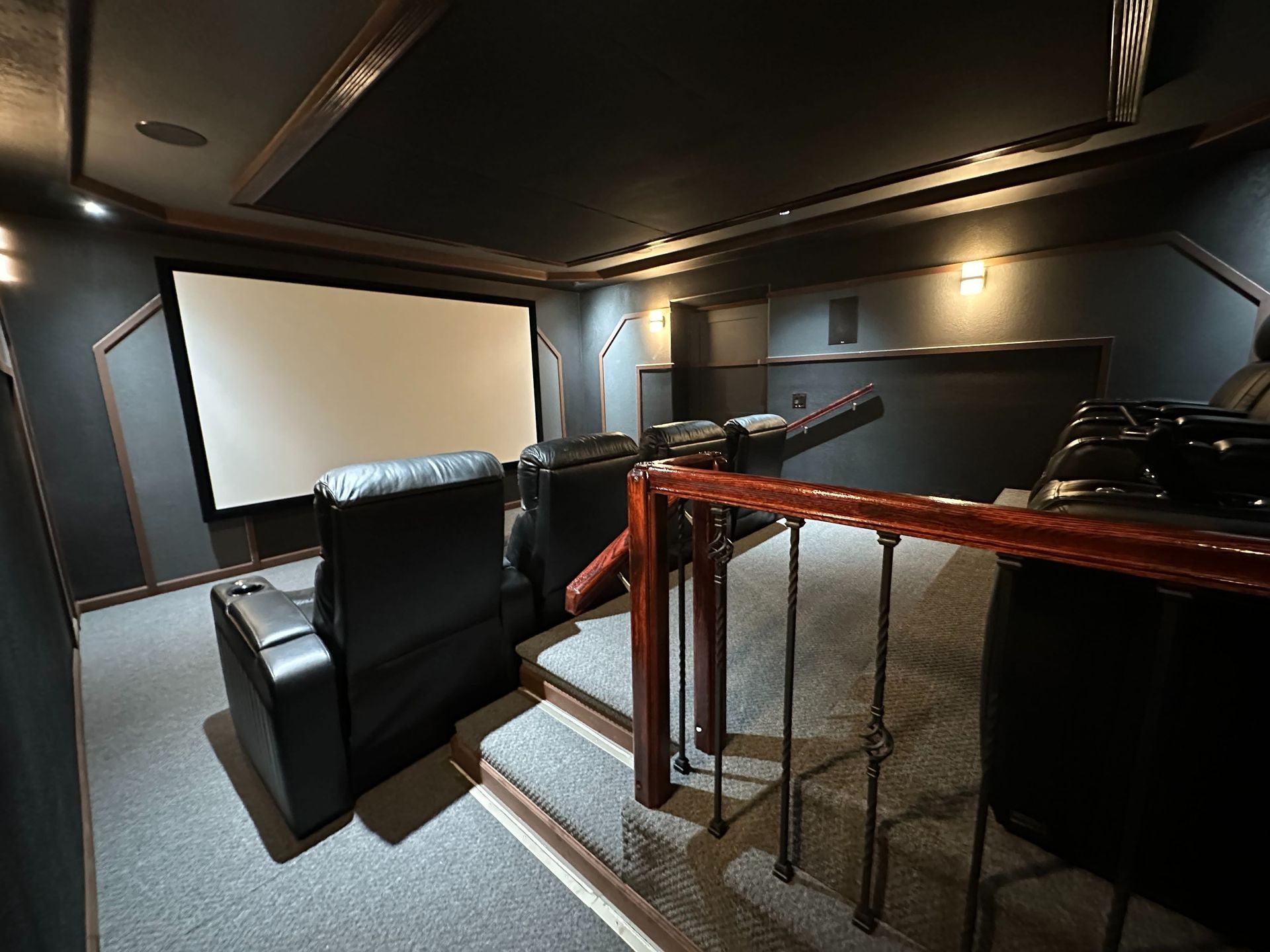 Dark home theater room with projector screen, black leather seats, and wood-railed stairs