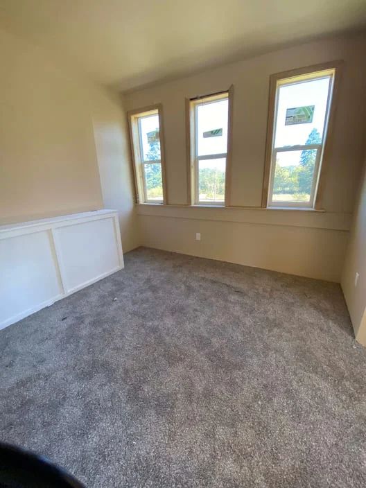Empty beige bedroom with three windows, gray carpet, and white wainscoting.